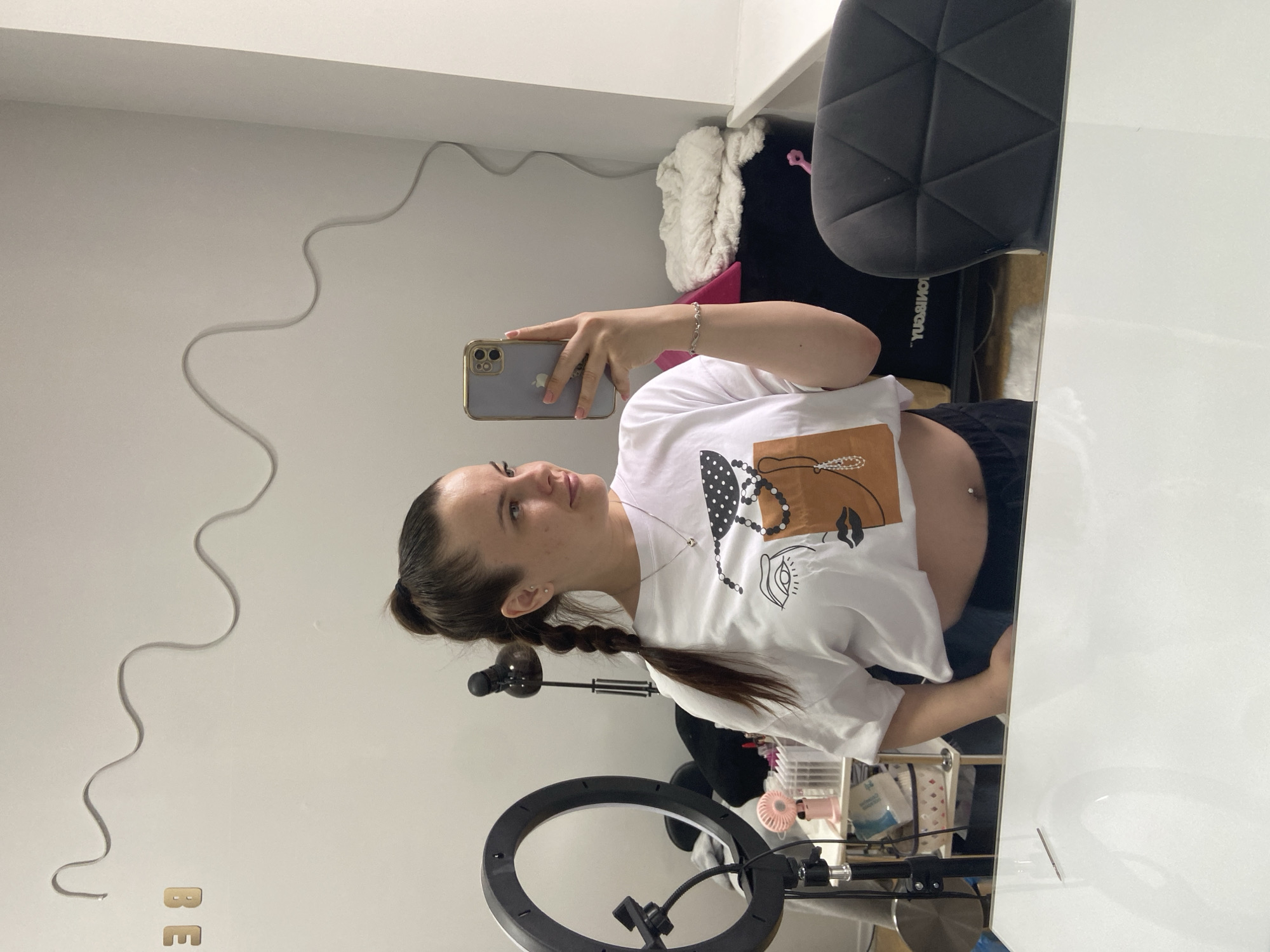 A medium shot captures a woman taking a mirror selfie in a brightly lit room. She is standing behind a white table, reflected in a mirror that covers most of the frame. The woman has brown hair pulled back in a high ponytail with a braid extending down her back. She is wearing a white t-shirt with a graphic print of a face and black drawstring pants, exposing her midriff where she has a navel piercing. Her left hand holds a gold-colored smartphone, angled to take the picture. A silver bracelet adorns her right wrist.
The background of the room includes a ring light on a stand to the left, a small pink fan on a shelf behind it, and various boxes and shelves filled with items. A black office chair with a geometric design is visible on the right side of the mirror's reflection, behind the woman. The wall behind her has a white wavy decorative element and the gold letters "BE" are on the upper left wall.
This scene is consistent with a private living space or a small studio setup, possibly for content creation. No specific landmarks or elements definitively place the location within Poltava, Ukraine.