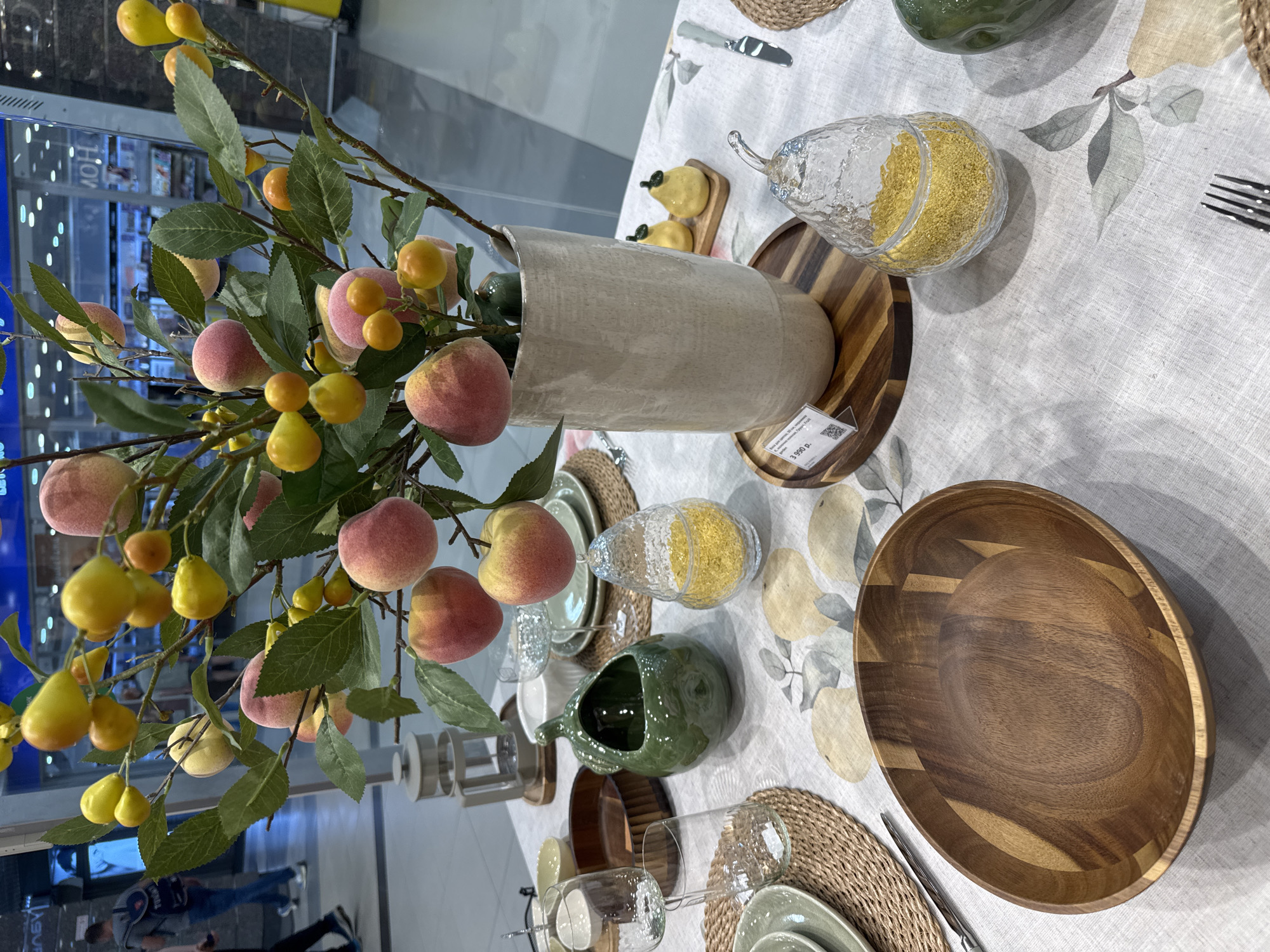 An overhead, slightly angled close-up shot captures a beautifully arranged table setting, likely a display in a retail store or showroom in Saint Petersburg, Russia.
The central feature is a tall, cream-colored, textured vase holding several artificial branches laden with faux peaches and smaller, bright yellow fruits (resembling small plums or pears) interspersed with green leaves.
The table is covered with a light-colored, possibly linen, tablecloth adorned with a subtle pattern of light grey and yellow fruits and leaves. The setting includes woven round placemats, stacked sage green ceramic plates, clear stemless glasses, and rustic wooden bowls with prominent grain, one large and one smaller that holds two miniature yellow ceramic fruits. A decorative clear glass jar, shaped like a pear and filled with bright yellow granules (possibly decorative sand or grains), sits on a wooden tray. A dark green, fruit-shaped ceramic jug or creamer is also part of the arrangement. Silverware, including a knife and a fork, is visible.
In the background, out-of-focus reflections on glass panels suggest an indoor commercial environment, with indistinct shapes of people and store fixtures visible. A white price tag is prominently displayed on a wooden tray, showing "3 990 р." (3,990 Russian Rubles) along with a QR code and smaller, undecipherable Russian text, likely describing the item (possibly "Поднос" for tray). The overall scene evokes a warm, autumnal, or harvest-themed home decor aesthetic.