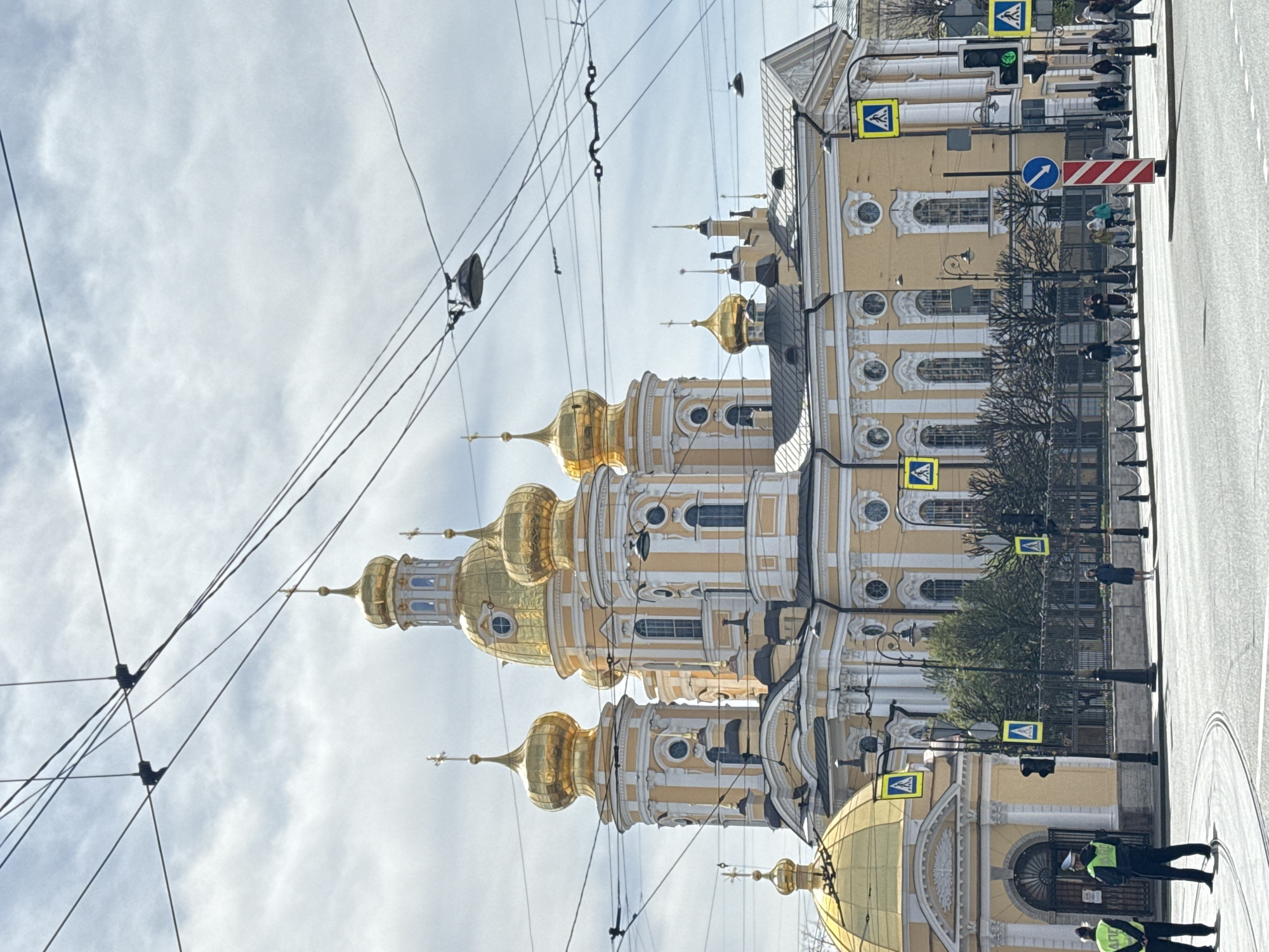 A daytime shot shows the Saint Andrew's Cathedral in Saint Petersburg, Russia. It has distinctive golden domes, and the building is yellow and white. There are power lines crisscrossing the upper portion of the image. The foreground includes part of a street and sidewalk, with pedestrian crossing signs. A few people are walking on the sidewalk.