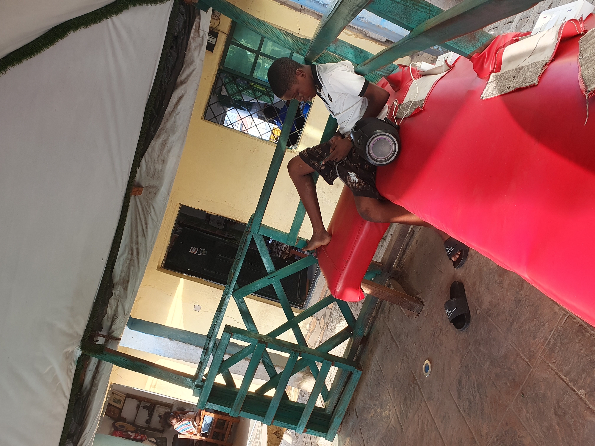 In Lokoja, Nigeria, a young boy is seated on a red padded bench outdoors, beneath a white canopy with green artificial grass trim. He is wearing a white polo shirt with a black collar and black and white patterned shorts. The boy is looking down at his phone, which he holds in his left hand. A small, black portable speaker is next to him on the bench. His feet are bare, and he is wearing black flip-flops. The bench is on a tiled patio. A green wooden railing is visible in the foreground. In the background, a yellow building with a dark green door and windows can be seen. A woman is seated in a wooden chair inside the building, near a shelf displaying pictures. The overall scene suggests a casual, outdoor gathering or recreational space.