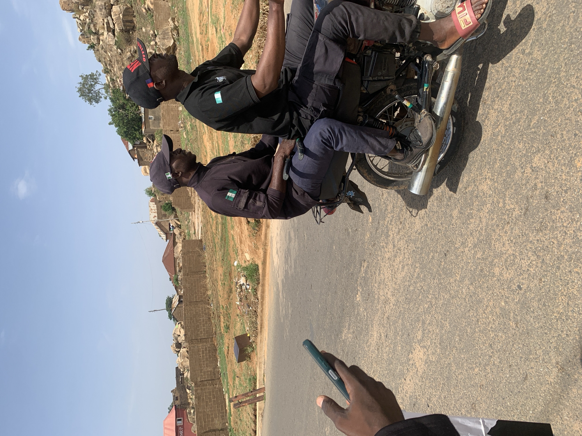Two men are seated on a motorcycle in Gura Top, Nigeria, alongside a road. The man in the front is wearing a black cap, black shirt, and dark pants. The man behind him is also wearing a black cap and black shirt. In the foreground, a hand holds a smartphone. The environment includes a paved road, rural buildings, vegetation, and a rocky hill in the background.