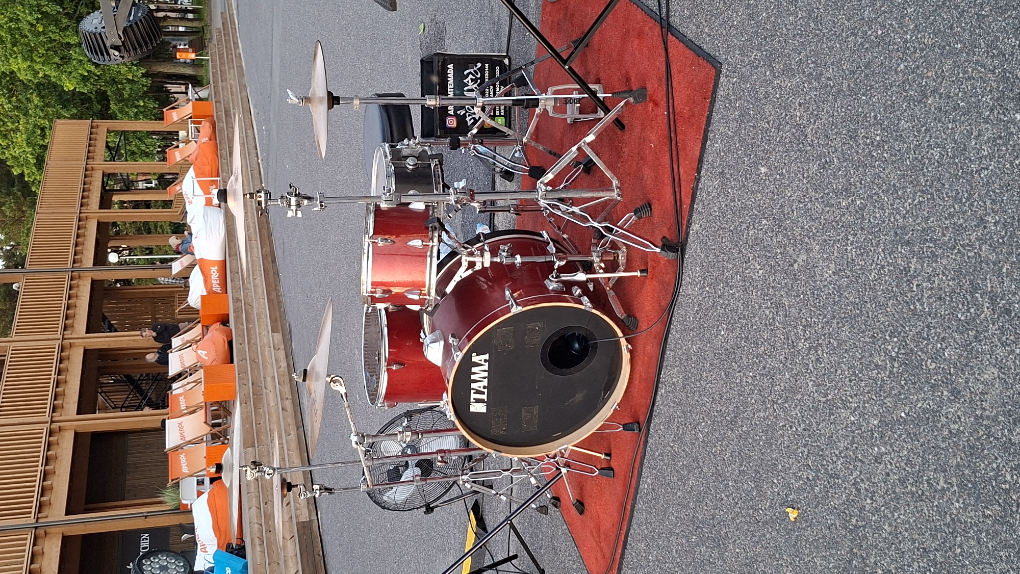 This is a media file of a red drum set on a red carpet placed on an asphalt surface. The drum set is the primary subject, with several individual drums and cymbals visible, all mounted on metallic stands. A black kick drum pedal is connected to the bass drum. A black box with white text is placed behind the drum set. To the left of the drums, a wooden structure with multiple levels and open sections is visible. Orange and white chairs are arranged on the wooden structure, and some people are seated in them. Trees and greenery are present behind the wooden structure. A black industrial fan is positioned in front of the drum set. Cables are visible on the carpet and extending onto the asphalt.
The image appears to be taken outdoors, possibly during the day, as there is ample light. The setting seems to be a casual outdoor venue or a performance space. The presence of people relaxing in chairs suggests a public gathering or event, perhaps a festival or an outdoor concert. The overall mood seems relaxed.
Visible text includes "APEROL" on the chairs, and on the black box behind the drum set, there is text that appears to be a logo with Korean characters and possibly contact information or a brand name. The word "TAMA" is visible on the bass drum head.