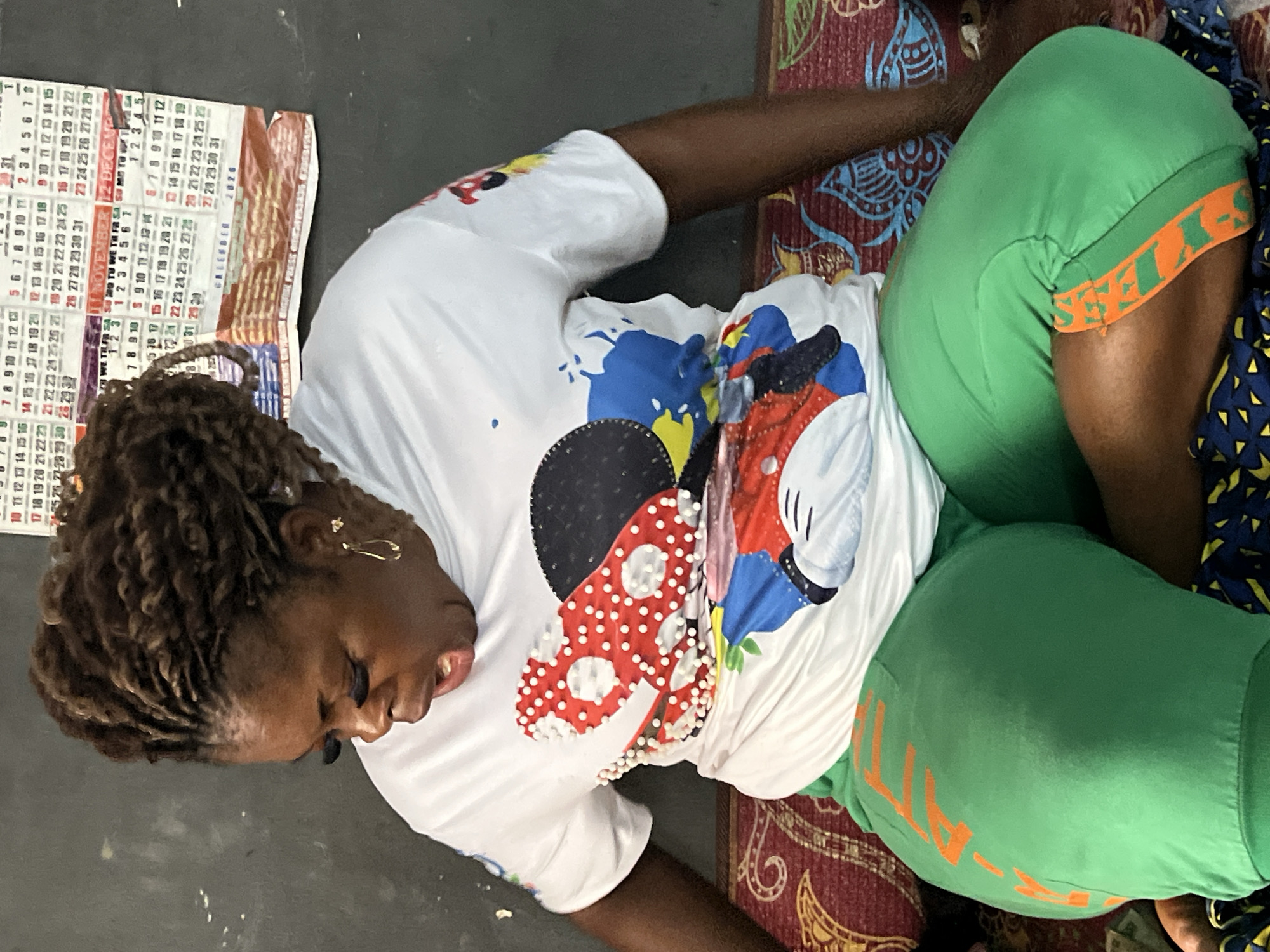 A Black woman with braided hair and dramatic false eyelashes is seated on a patterned couch in what appears to be a room. She is wearing a white t-shirt with a colorful Mickey Mouse graphic and bright green pants with orange lettering. The woman's mouth is open, and she is looking down. In the background, a calendar is visible on a dark wall. The overall impression is of a casual, indoor setting, possibly a home or a small business, located in Nkpor, Nigeria.