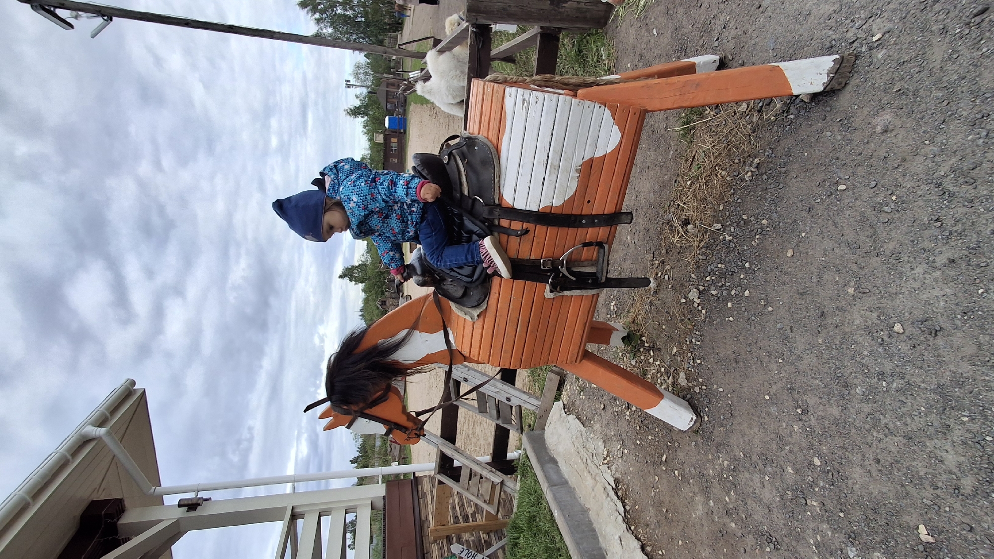 In the location of Лукаши, Russia, a young child wearing a blue hat and patterned jacket is seated on a wooden horse ride. The horse is painted orange and white with a black mane. It has a saddle and stirrups. The ground around the horse is gravelly. There is a white building with a brown trim in the background. The sky is overcast.