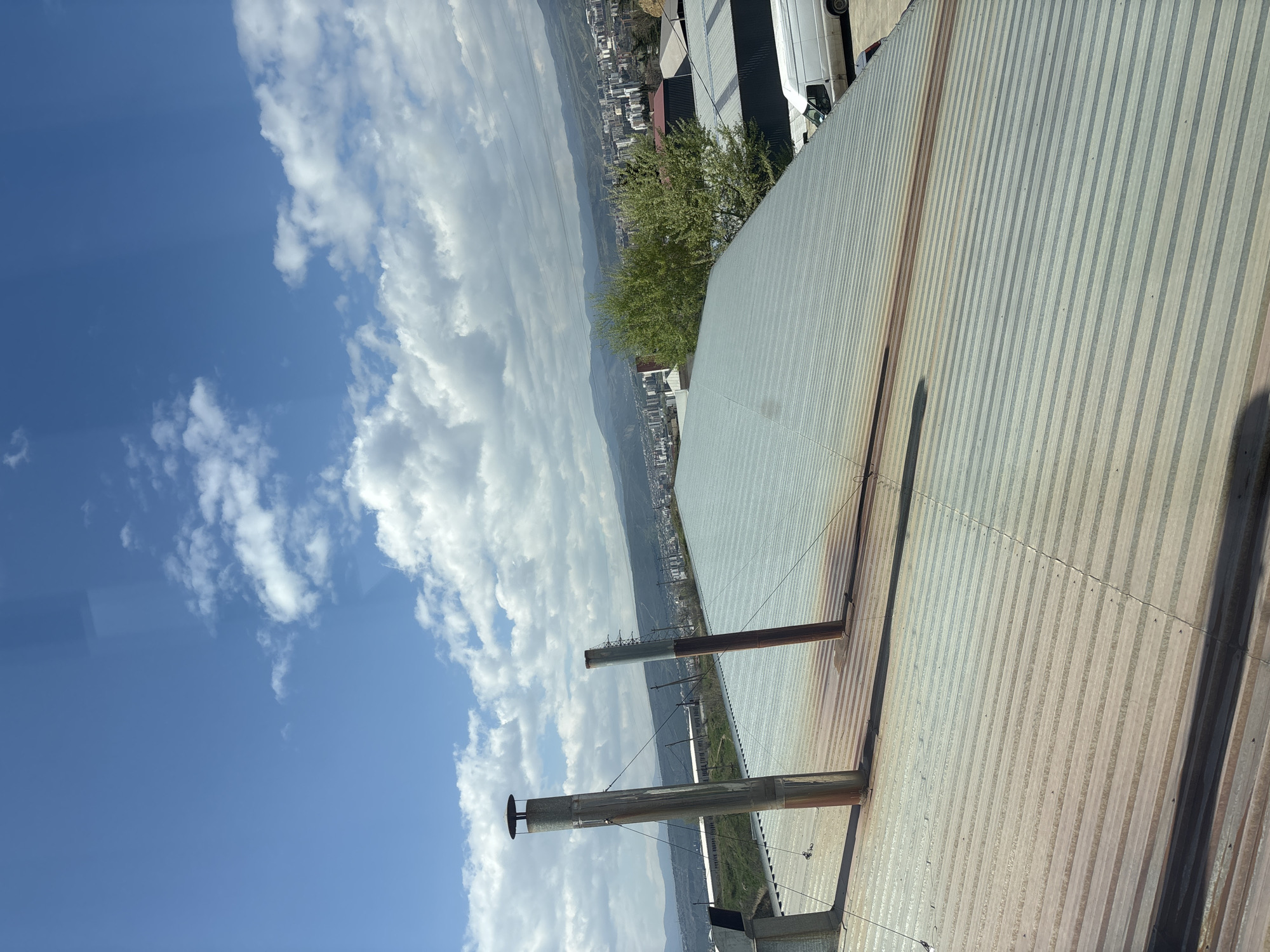 The image presents a scene above a corrugated metal roof, likely of an industrial or commercial building in Tbilisi, Georgia. The roof has several ventilation pipes or chimneys extending upwards. In the background, a cityscape with buildings and mountains is visible under a sky of blue and scattered white clouds. A couple of white vehicles are parked at the roof's edge.