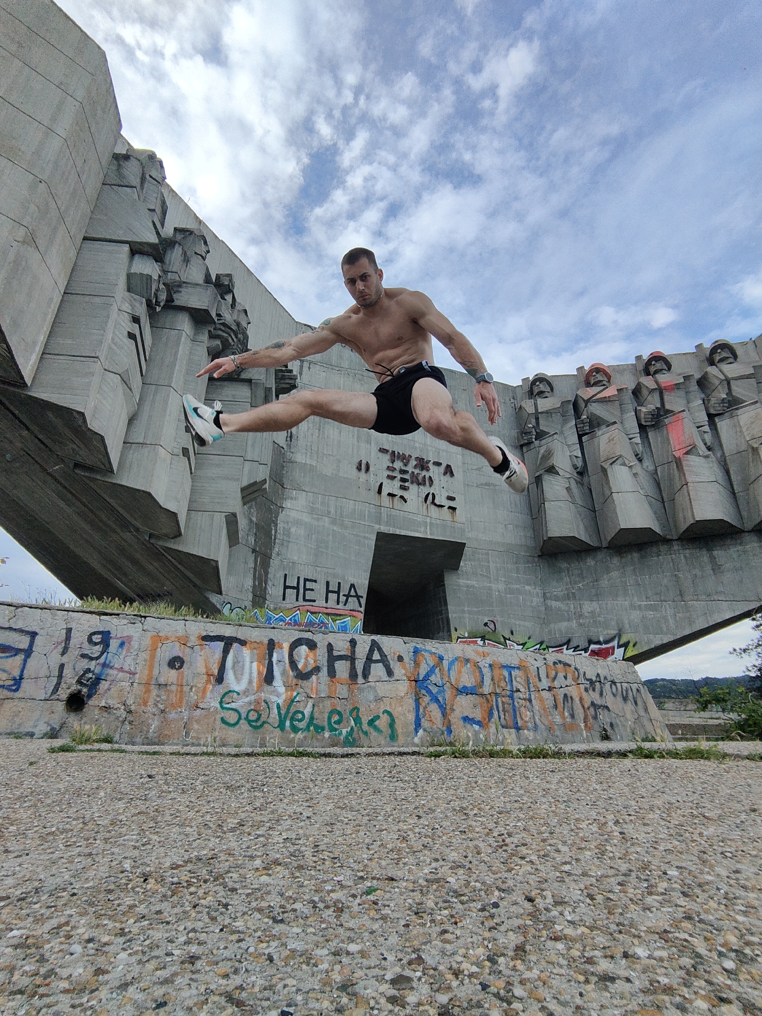 A man is captured mid-air during a dynamic jump, positioned centrally against a monumental backdrop. He is shirtless, wearing black shorts, white sneakers with turquoise accents, and a watch on his left wrist. His legs are extended in a wide split, and his arms are outstretched, with his left hand pointing forward, conveying an athletic or parkour-like movement. He has a focused expression, and tattoos are visible on his arms.
The background features a large, Brutalist-style concrete monument, characteristic of Soviet-era design, located in Varna, Bulgaria. The monument consists of imposing, abstract block figures that resemble soldiers or workers, rising sharply towards the sky. Some of these figures have faint red accents on what appear to be helmets or shoulders. The concrete surface of the monument shows signs of age and graffiti, with Cyrillic text visible, notably "HE HA" and "ЖЪЛТА СЕКИ" (Yellow Axe/Cut) spray-painted on its lower sections.
In the foreground, a lower concrete wall is heavily covered in colorful graffiti, including the prominent text "TICHА" in black and "Severler" in green, along with other scribbled numbers like "19". The ground surface beneath the wall is a coarse, pebbled concrete. The scene is set outdoors during daytime, under a partly cloudy sky, with ample natural light. The man's active and vibrant pose contrasts with the austere and somewhat weathered appearance of the monument.