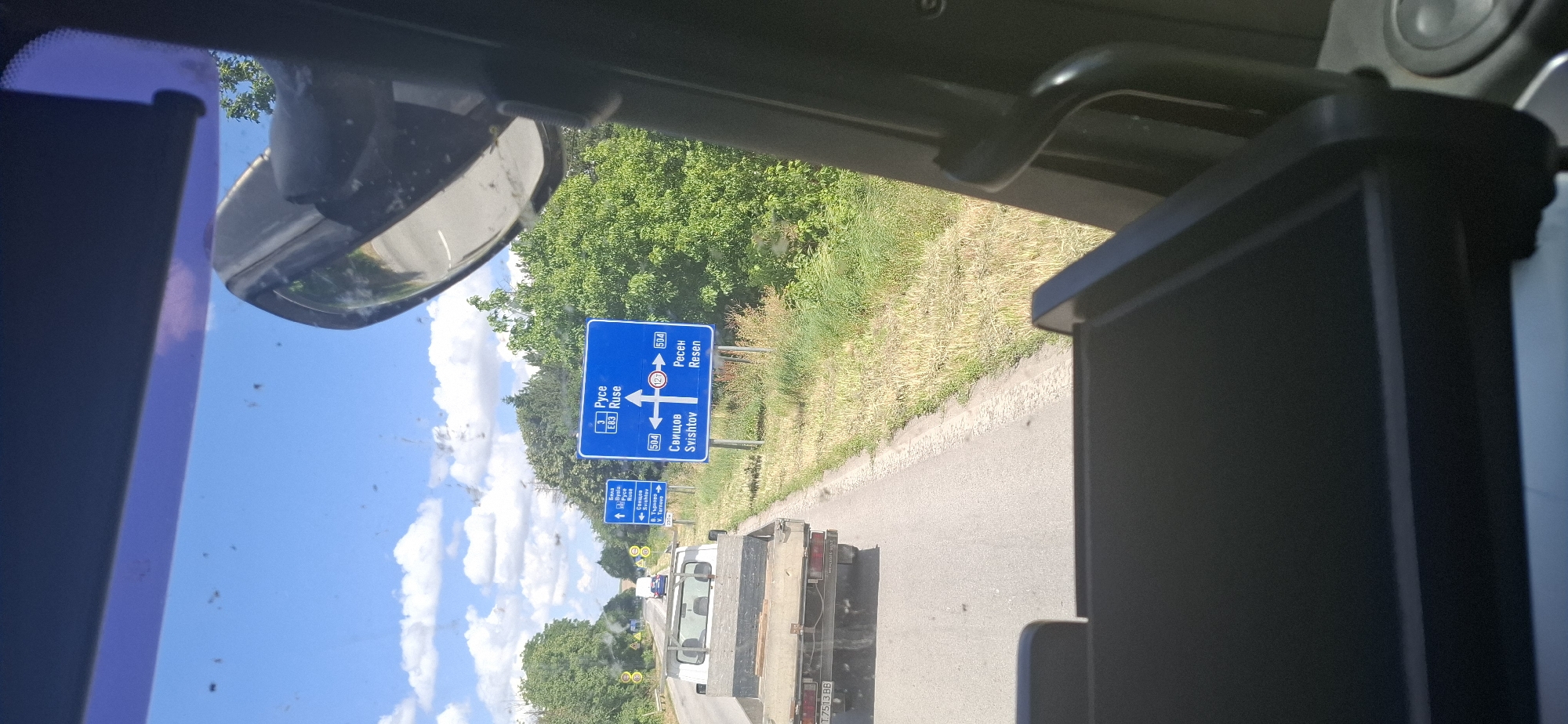 The image shows a road scene in Maslarevo, Bulgaria, on a sunny day. A blue road sign indicates directions to Ruse (3, E83), Svishtov (504), and Resen (504). There is also a smaller blue sign with a white arrow pointing left, indicating directions to Byala (3), and Veliko Tarnovo (2). In the foreground, a white flatbed truck is visible from behind, with the license plate T 7513 BB. Another vehicle is partially visible further down the road. The surrounding environment is characterized by green trees and dry grass. The perspective is from inside a vehicle, likely a truck, with a side mirror and part of the dashboard visible in the frame. The sky is blue with scattered white clouds.