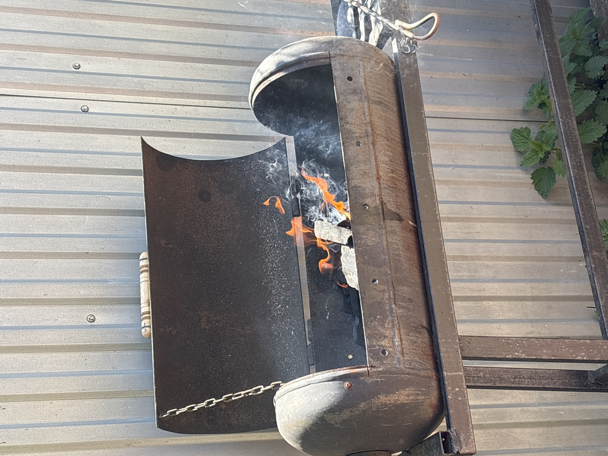 A metal fire pit or smoker is affixed to a corrugated metal wall. Flames and smoke are visible within the pit, which has an open door attached by a chain. The pit appears to be made from repurposed metal tanks. The wall and fire pit are outdoors, with vegetation visible in the upper right corner. There is no confirmation this image was taken in Prikamskii, Russia, and the location is not discernible.