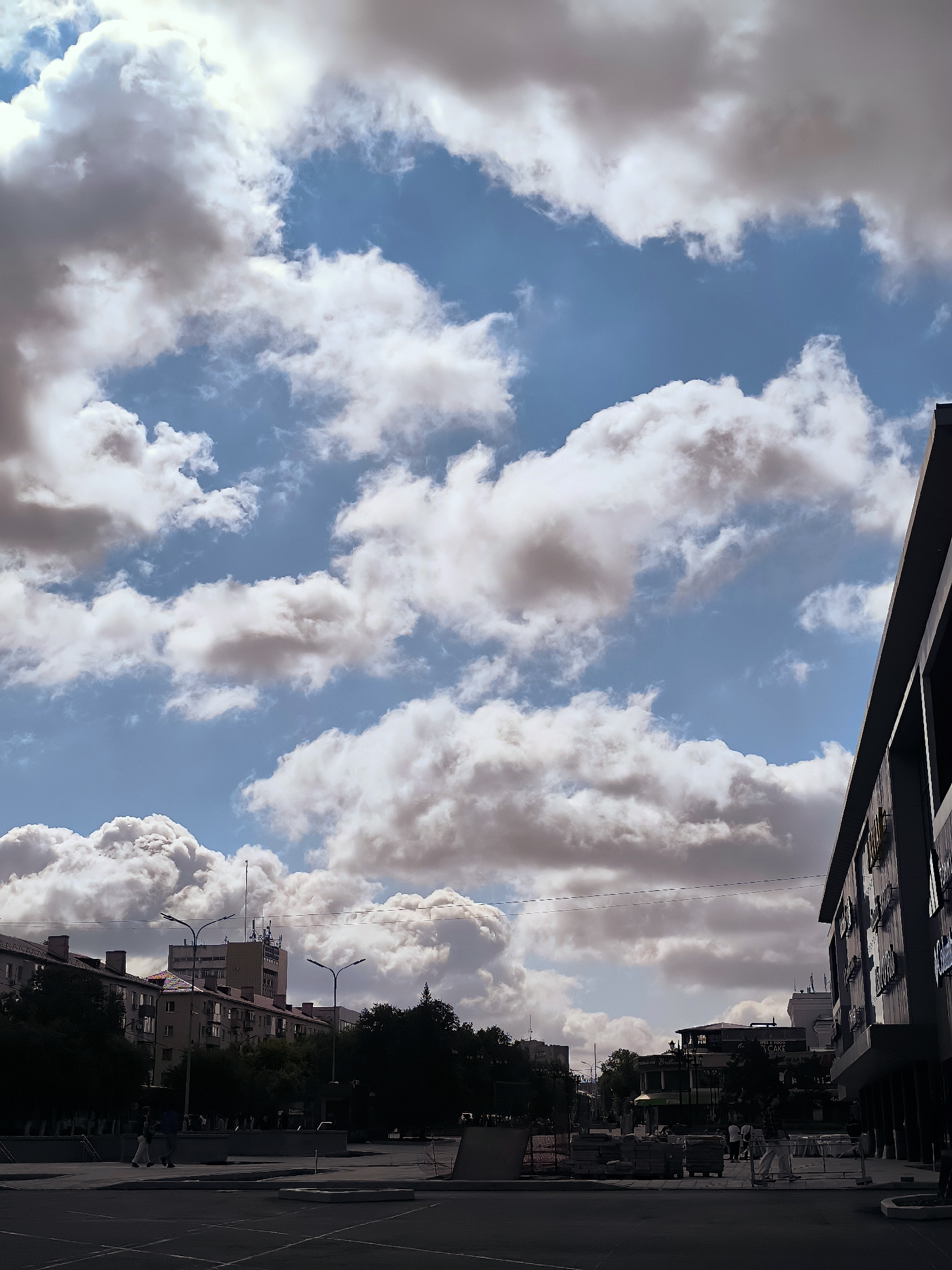 The image depicts an urban scene under a partly cloudy sky in Qarağandı city administration, Kazakhstan. In the foreground, a dark asphalt surface, possibly a street or plaza, is visible with some markings and concrete structures. To the right, a modern building with dark facade and large windows extends into the frame. In the middle ground, a street with parked cars and pedestrian walkways is lined with buildings and trees. The buildings are of varying heights and architectural styles, with some appearing to be residential apartments and others commercial establishments. A tall building with a tower and antennas stands in the background, possibly a government or office building. A prominent feature of the sky is the presence of large, fluffy white and gray clouds interspersed with patches of blue sky. A few people are visible walking on the sidewalks and crossing the street. The overall impression is a typical urban environment on a day with dynamic cloud formations.