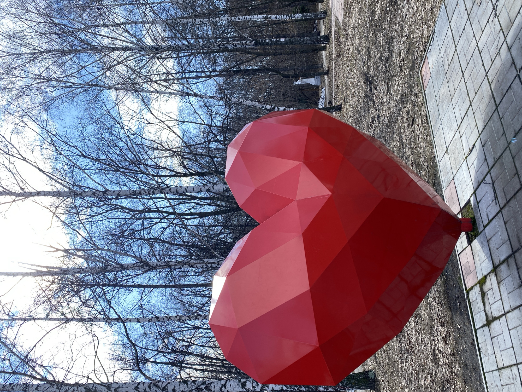 The image shows a large, geometric red heart sculpture in a park setting, likely in Ulyanovsk, Russia. The sculpture sits on a concrete tile surface that transitions to a dirt and leaf-covered ground. Behind the sculpture, a grove of birch trees with bare branches stretches toward a blue sky with scattered clouds. The picture is taken from a low angle, tilting upwards. The lighting appears bright and sunny, casting some shadows.