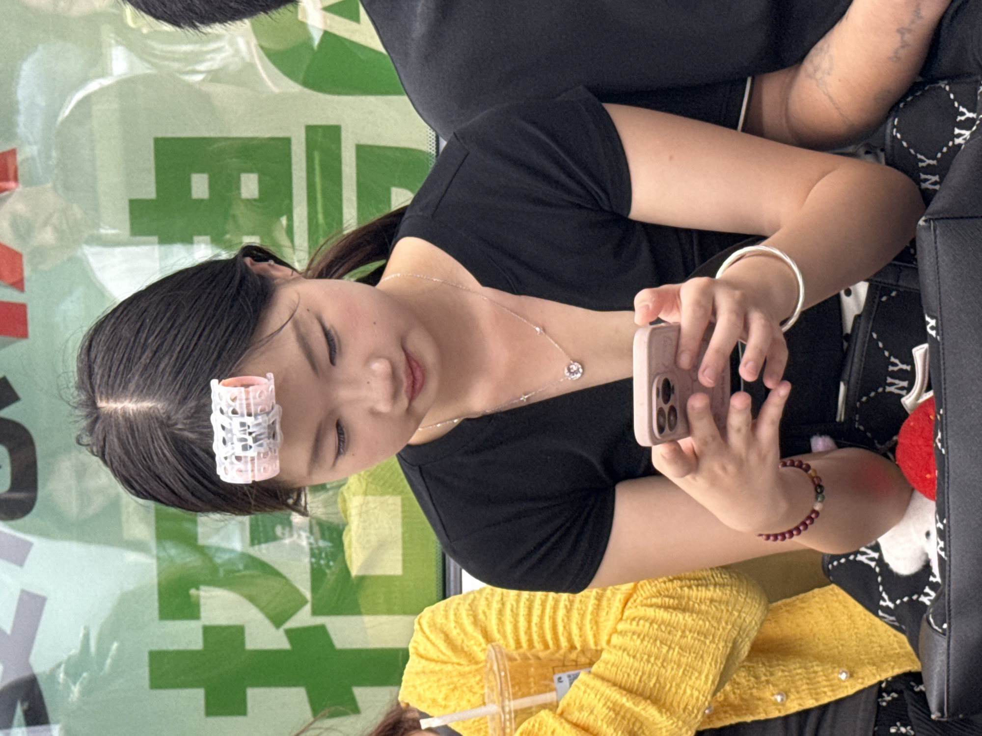 A young woman with a hair roller is seated, looking at a mobile phone in her hand. She wears a black shirt, a silver necklace, and bracelets on both wrists. Part of a yellow, textured shirt and a beverage cup with a straw are also visible. The background includes an advertising sign in green and white with what appear to be Chinese characters, suggesting a location in Yuzhong District, China.