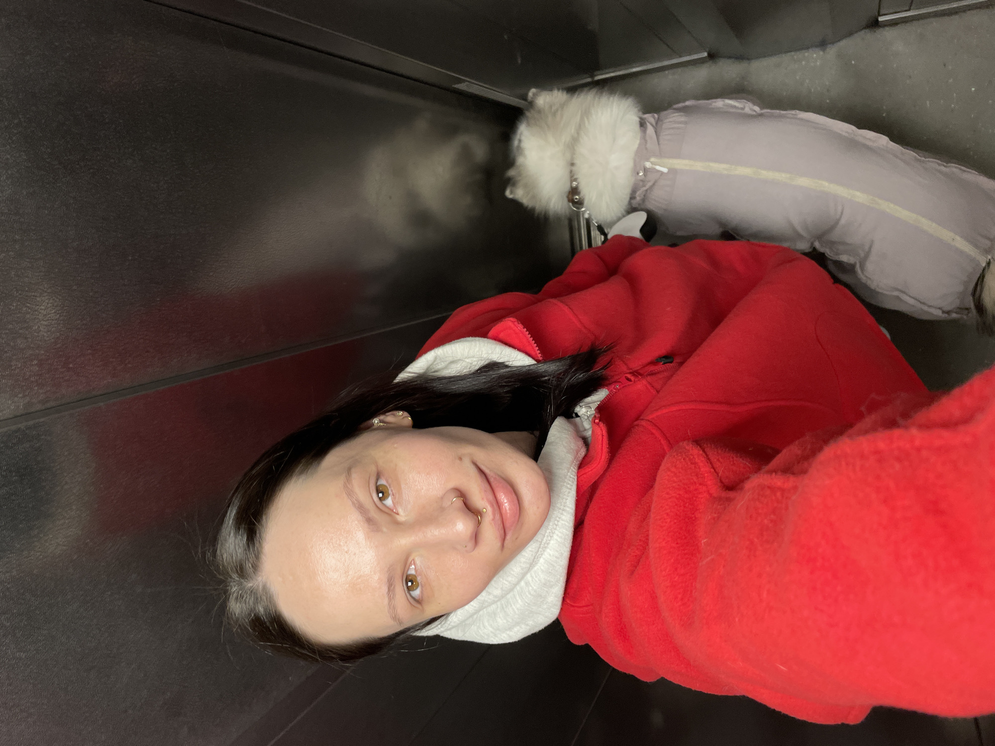 The image shows a woman and a dog indoors, likely in an elevator, though Izhevsk, Russia's location cannot be verified from the image. The woman has dark hair, fair skin, and facial piercings. She's wearing a red hooded jacket. Beside her, a dog is wearing a grey coat with a white fur trim. The background is a metal surface, indicating the elevator. The image appears to be a selfie.