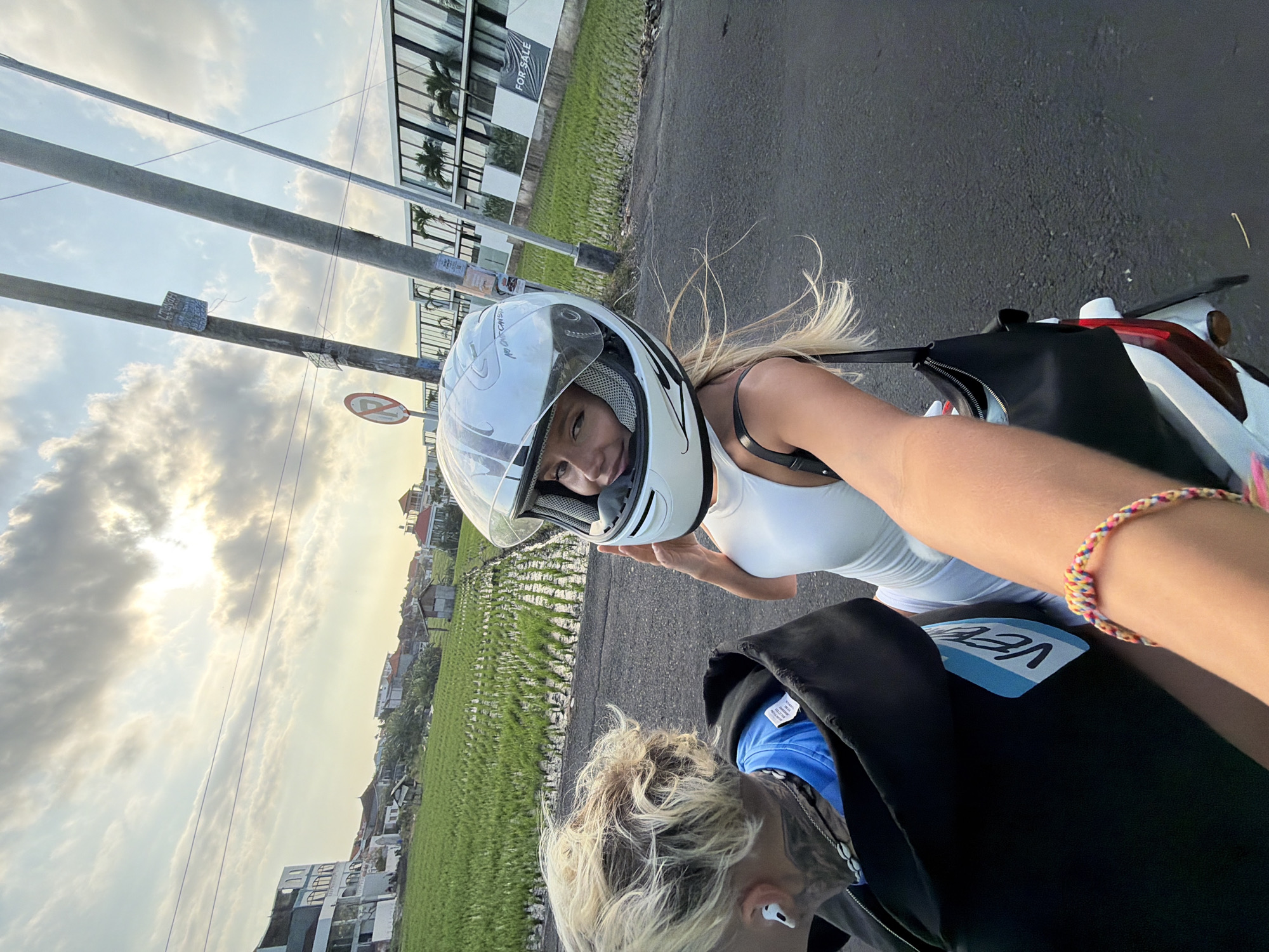 Two individuals are on a motorcycle, viewed from the perspective of the driver, who is taking a selfie. The passenger, wearing a white helmet, smiles at the camera. She has blonde hair and is wearing a white sleeveless top. The driver, in the foreground, has blonde hair and is wearing a black hooded jacket over a blue shirt. A portion of their arm, adorned with a colorful braided bracelet, is visible. They are on a paved road with a rice paddy field to the left, bordered by buildings. To the right of the road, utility poles and a modern building with "FOR SALE" signage are visible. The sky is overcast with sunlight breaking through the clouds. The scene appears to be in North Kuta, Indonesia, given the presence of rice paddies and tropical vegetation.