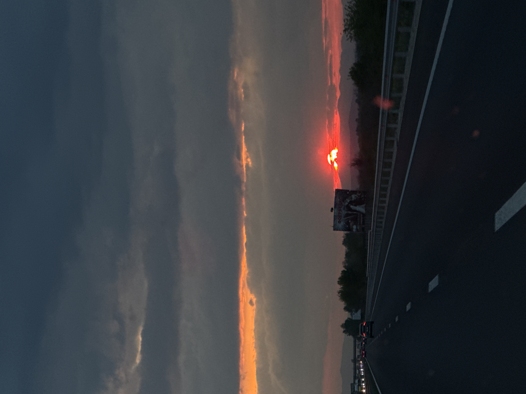 The image captures a dramatic sunset or sunrise over a highway in Malo Konare, Bulgaria. The sky, when viewed in its correct horizontal orientation, dominates the upper portion, displaying a striking contrast between dark, heavy blue-grey clouds and a vibrant, intense orange-red sun positioned towards the right horizon. The sun's powerful light illuminates a long, fiery streak within the clouds, extending across the mid-sky.
The lower part of the frame features a dark asphalt highway, stretching into the distance with clear white lane markings. A metal guardrail runs alongside the road, separating it from a dense strip of dark trees and foliage. Beyond the trees, the silhouettes of distant mountains or hills are visible against the reddish glow of the horizon. Several cars can be seen further down the highway, identifiable by their illuminated red taillights.
A large billboard with partially legible Cyrillic text stands on the right side, above the trees and guardrail. The time of day is clearly either dusk or dawn, given the intense colors of the sky and the low-lying sun. The scene conveys a sense of travel and the natural beauty of the Bulgarian landscape at a specific, dramatic moment.