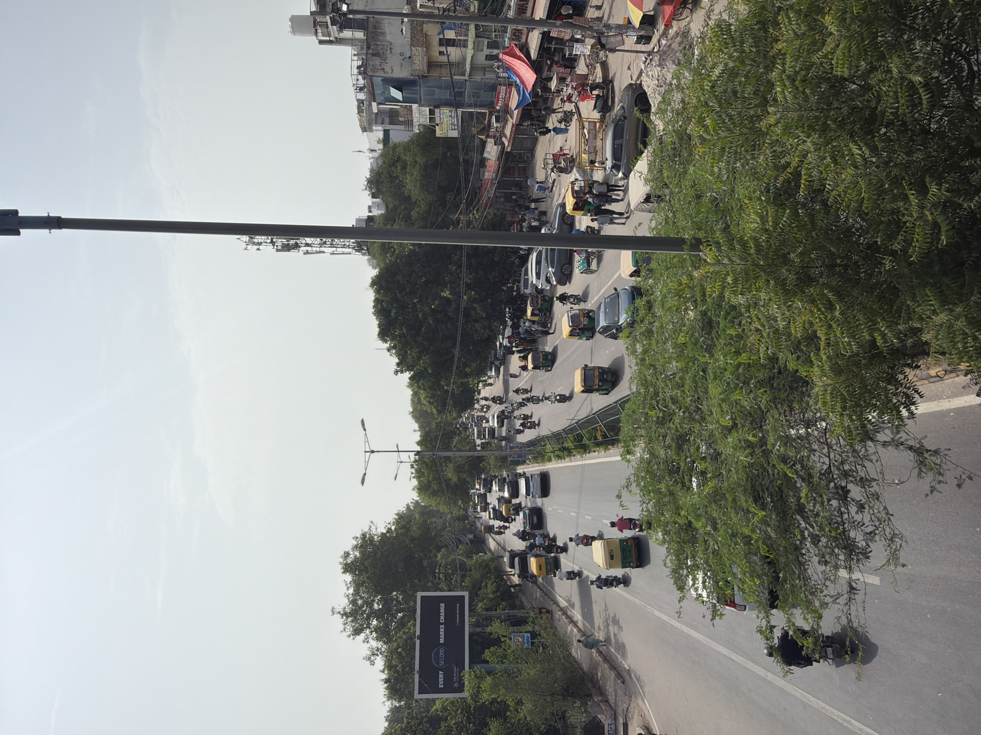 The image depicts a busy street scene in Delhi, India, likely during daylight hours. A multi-lane road is filled with traffic, primarily consisting of auto-rickshaws, motorcycles, cars, and a few pedestrians. The vehicles are moving in both directions, with some congestion visible. On the left side of the road, a billboard displays the text "EVERY SECOND MARKS CHANGE". Trees line the road, and buildings of varying heights and architectural styles are visible on the right side, suggesting an urban environment. Several power lines and a tall telecommunications tower are also present in the scene. The overall atmosphere appears to be that of a typical bustling day in a crowded Indian city.