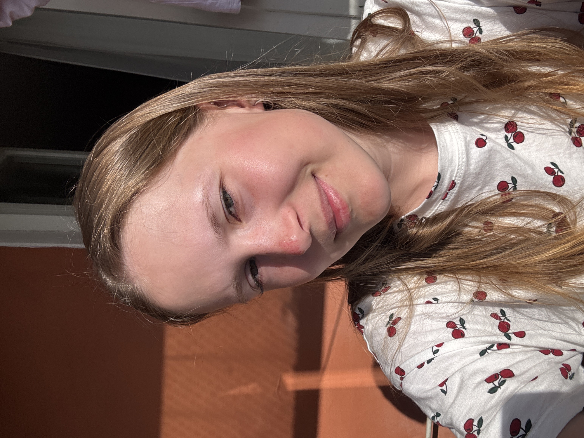 A young woman with blonde hair is wearing a white shirt with a red cherry print. She is outdoors, in front of an orange wall and a window. The location is not specified, so determining if it's Chaikovsky, Russia, is not possible.