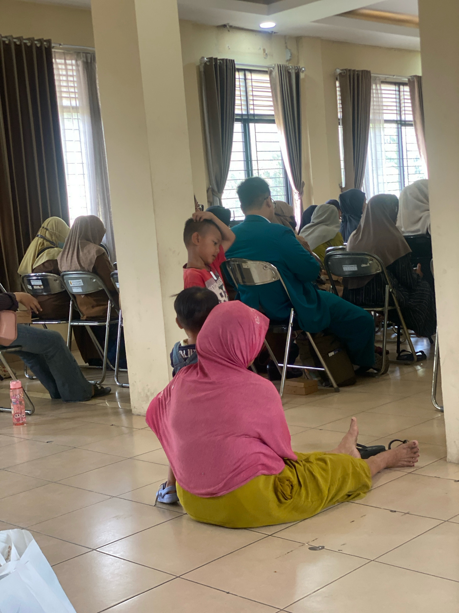 The image depicts an indoor scene, likely a community hall or waiting area, with several people seated in chairs and on the floor. In the foreground, a person wearing a pink headscarf and a yellow skirt is seated on the tiled floor, facing away from the camera. To their left, a water bottle with cartoon characters is visible on the floor.
Further back, a young boy in a red shirt is standing and looking to his right, with his hand on his head. Behind him, another child is partially visible. Several other individuals, mostly women wearing headscarves, are seated in rows of folding chairs. Some are looking forward, while others are looking to the side or down. The room has large windows with blinds and brown curtains, and a pillar is prominently visible in the center of the image. The lighting suggests it is daytime, with natural light coming from the windows and overhead fixtures.
The scene appears to be from a public gathering or an event where people are waiting, possibly for services or a program. The location is identified as the city of Banjarbaru, Indonesia, which suggests this is a local community event. The overall atmosphere is one of a quiet, orderly gathering.