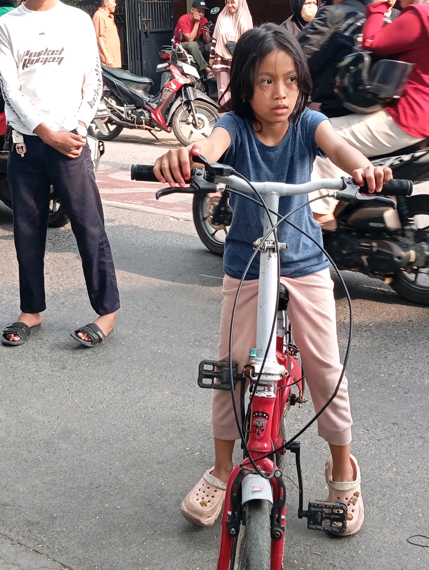 In an outdoor urban setting, likely a street in Bandung City, Indonesia, a young girl is the central subject, posed on a red bicycle. She has dark hair and is wearing a blue t-shirt and light pink pants. Her gaze is directed slightly to her right. The bicycle she is on appears to be a children's model, with visible handlebars, brakes, and pedals.
In the background, other individuals and vehicles are present, suggesting a public area with activity. To the left, a person in a white long-sleeved shirt and dark pants stands with their hands clasped. Behind them, motorcycles are visible, one with a prominent red and black design. Further back, other people are seen, including one wearing a red shirt and a black cap, and others in more indistinct clothing. The ground is paved with asphalt, and a hint of a building or fence can be seen in the background. The lighting suggests it is daytime.