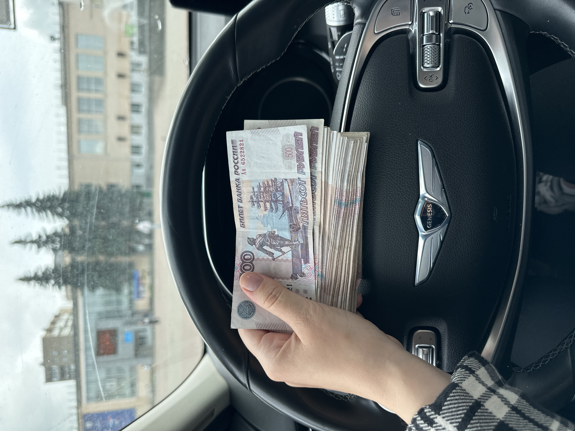 A person's hand, with manicured fingernails, holds a thick stack of Russian rubles in front of a car's steering wheel. The currency is predominantly 500 ruble notes, visible with images of ships and text. The steering wheel has a "GENESIS" emblem in its center, and various control buttons are visible. The background, seen through the car's windshield, shows a street scene with trees, buildings, and a fountain. The sky is overcast. The location is identified as Chaikovsky, Russia.