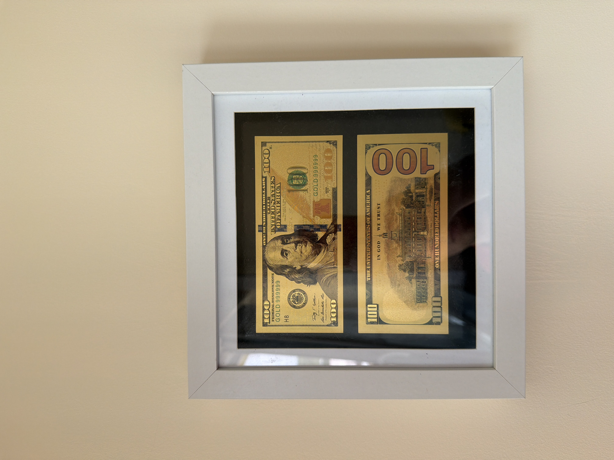 A white square picture frame hangs on a plain, light beige wall. Inside the frame, two golden novelty hundred-dollar bills are displayed side-by-side against a black mat.
The bill on the left represents the front of a hundred-dollar bill, featuring a golden rendition of Benjamin Franklin's portrait. Visible text includes "100" (appearing multiple times), "FEDERAL RESERVE NOTE," "GOLD 999999" written vertically, "ONE HUNDRED DOLLARS," and "H8" as a serial letter/number. Parts of "THE UNITED STATES OF AMERICA" are also visible.
The bill on the right depicts the reverse side, also in gold, showing the Independence Hall. Text legible on this bill includes "100" (with the large numeral appearing as "OOL" due to its orientation), "THE UNITED STATES OF AMERICA," "IN GOD WE TRUST," and "ONE HUNDRED DOLLARS." "GOLD 999999" is partly visible.
The setting is indoors, with bright, even lighting illuminating the display. No activity is taking place other than the static display of the framed items. The image itself does not provide any specific cues regarding the time of day, weather, or exact location within Chervena voda, Bulgaria, beyond being a well-lit interior.