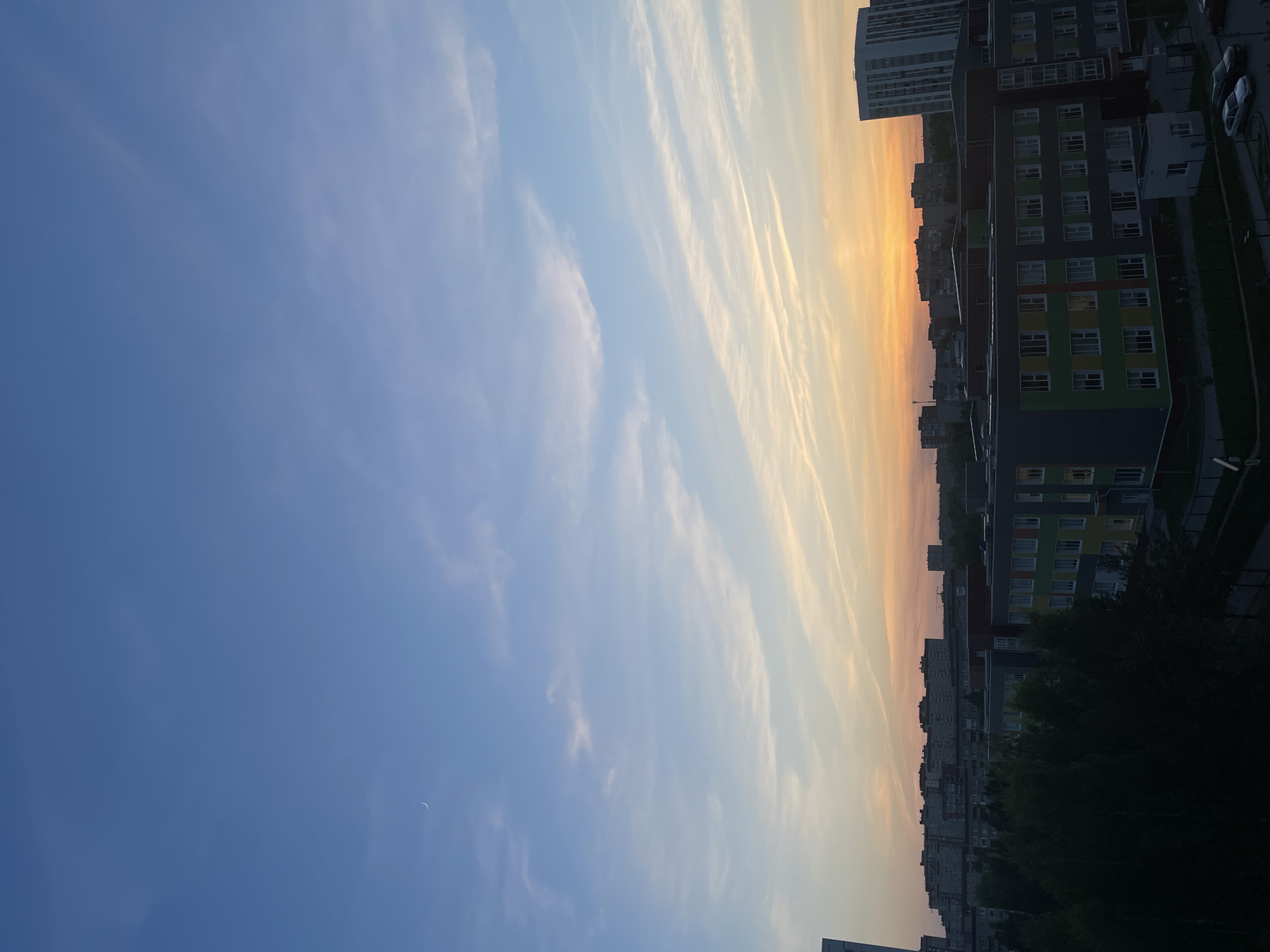 The image shows a sunrise or sunset in Izhevsk, Russia. The sky is primarily blue, fading to orange and yellow near the horizon, with thin, streaky clouds. Tall apartment buildings dominate the right side of the frame, some with colored panels. Trees are visible in front of the buildings. Some parked cars are visible between the buildings. The buildings appear to be Soviet-era apartment blocks.