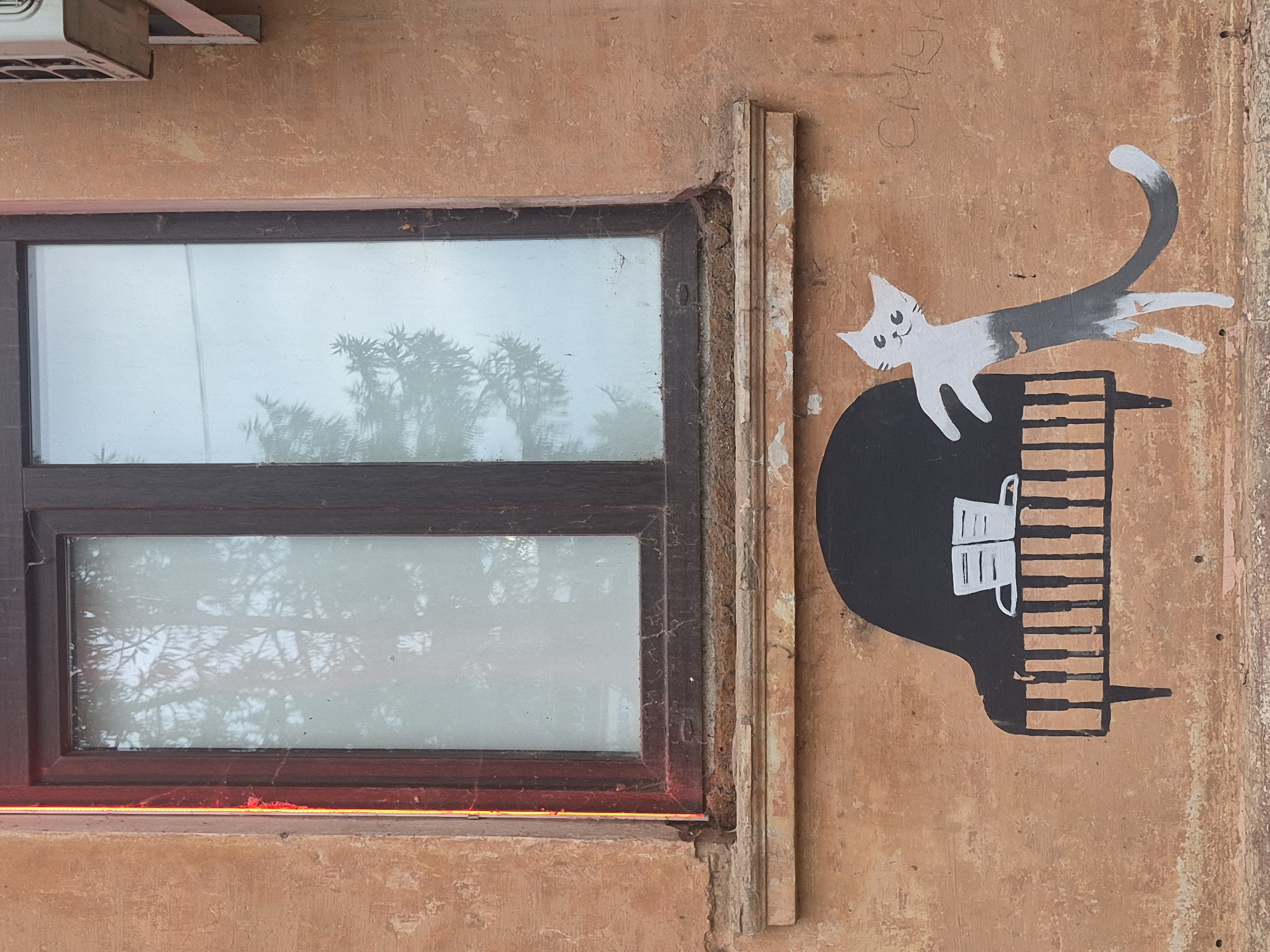 This is an exterior shot of a building in Votkinsk, Russia. The building has an orange stucco facade and a dark brown wooden window frame. The window is double-paned and reflects trees. Below the window, on the facade, is a mural depicting a black grand piano with white keys. Two white books sit on top of the piano. To the right of the piano is a stylized white cat with gray markings. The cat appears to be dancing or playing the piano. The cat's body is elongated and curves to the right, with its tail extending horizontally. The cat has a smiling face with large black eyes. To the right of the mural, there is faint graffiti on the wall. An air conditioning unit is visible on the upper right portion of the building. The lower part of the facade shows signs of wear and tear, with chipped paint and some debris at the bottom.