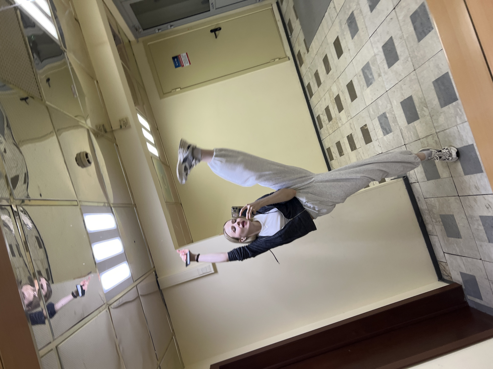 The image depicts a young woman in a mirror-lined hallway in Khimki, Russia. She is wearing gray sweatpants, a white t-shirt, and a dark zip-up jacket. She is holding a smartphone and appears to be taking a selfie. The hallway has a tiled floor with a gray and white pattern, and yellow walls. A door is visible at the end of the hallway.