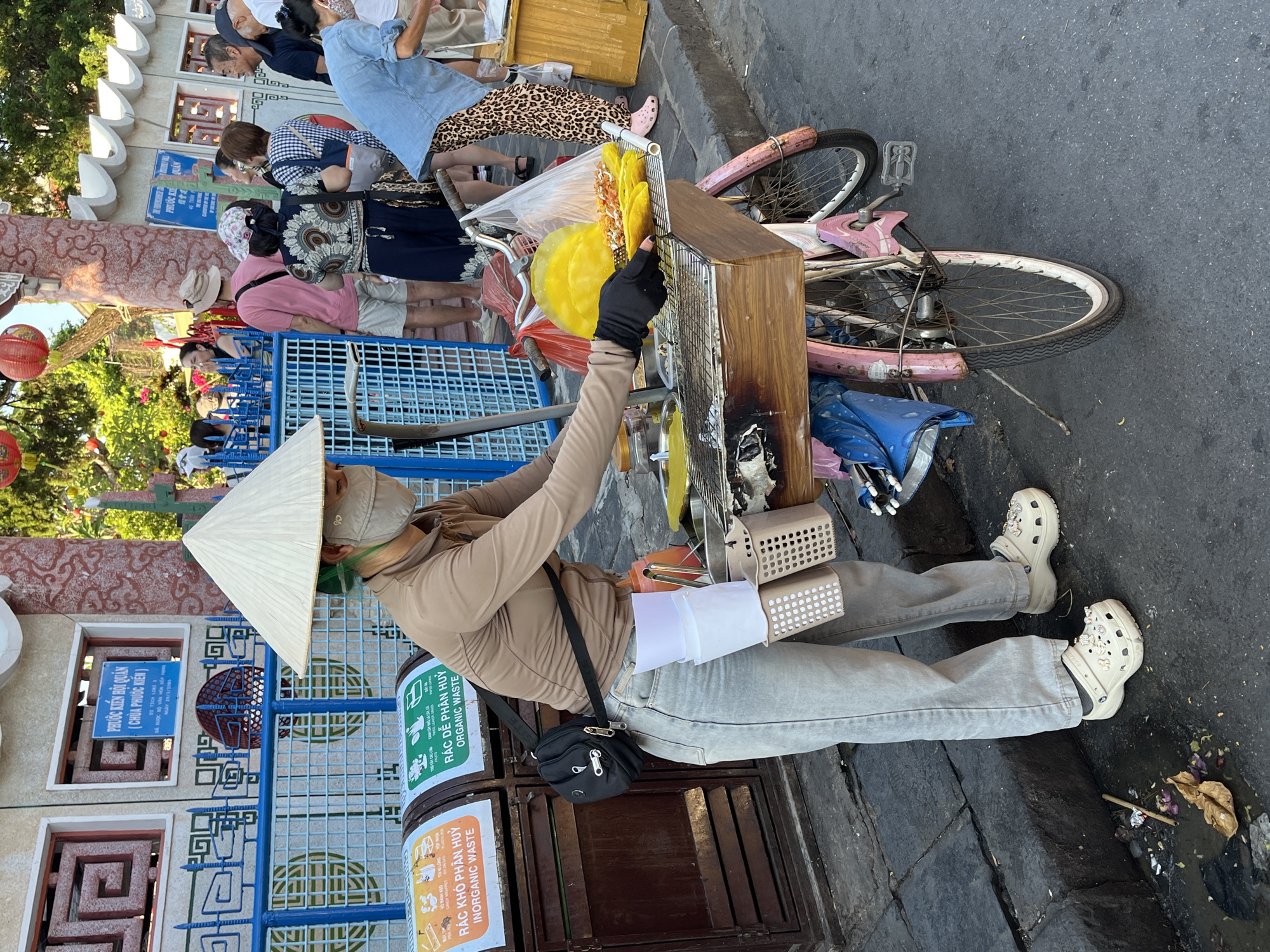 A street vendor wearing a tan outfit, a white conical hat, a face mask, and Crocs sits alongside a pink bicycle-powered stand in Hội An Ward, Vietnam. The vendor is preparing a yellow food item, possibly pancakes, wearing black gloves. The bicycle stand is cluttered with items, including yellow food, a wooden box, and various containers. Nearby are trash cans labeled "Rác Dễ Phân Hủy Organic Waste" and "Rác Khó Phân Hủy Inorganic Waste". In the background, tourists are seen near a building with a sign that reads "Phước Kiến Hội Quán (Chùa Phước Kiến)" indicating a historical site. The area is likely a pedestrian zone.