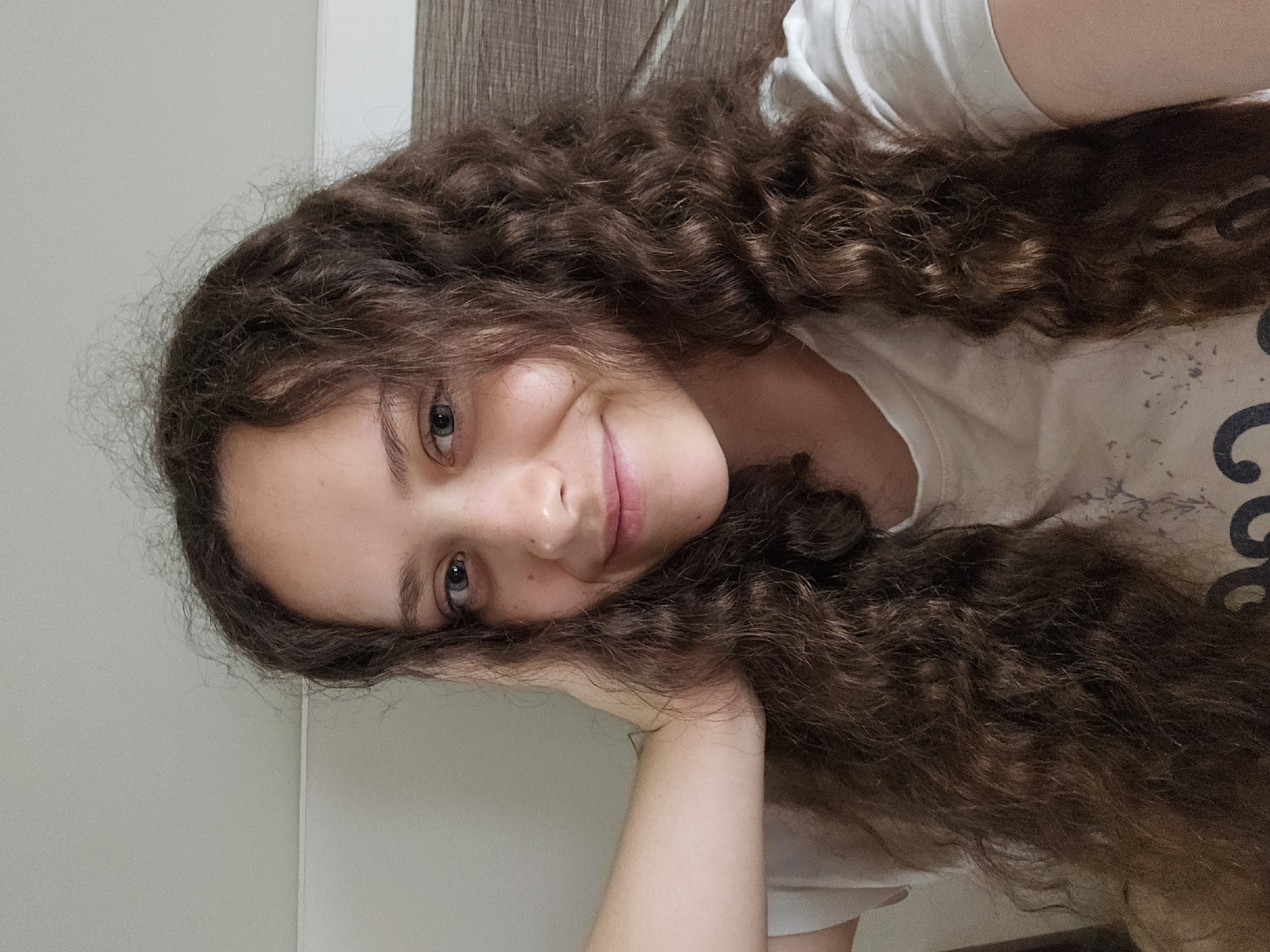 A young woman with long, curly brown hair and blue eyes is shown from the chest up, in a close-up selfie. She is wearing a white t-shirt. Her right hand is resting on the back of her head, and she is smiling gently at the camera. The background is a plain, neutral color, with a hint of wood grain texture to the right. The lighting is soft and natural, creating a warm and relaxed atmosphere.