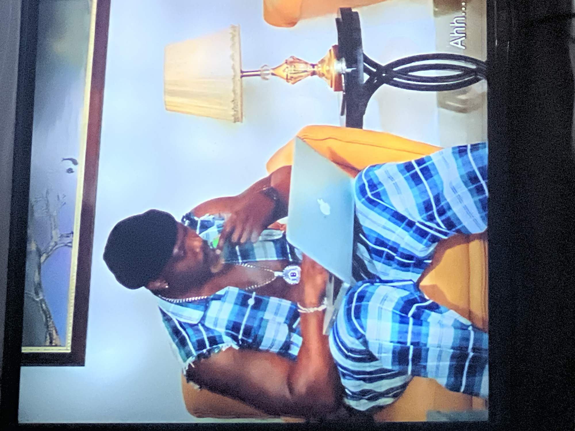 A man is sitting in a chair and working on a laptop. The man is wearing a plaid shirt and pants, and a black beanie. He has a necklace with a pendant that has the letter "B" on it. There is a lamp on a table next to the chair. The background is a wall with a framed picture on it. The text "Ahh..." is visible in the lower right corner of the image. This image is not in Lagos, Nigeria.