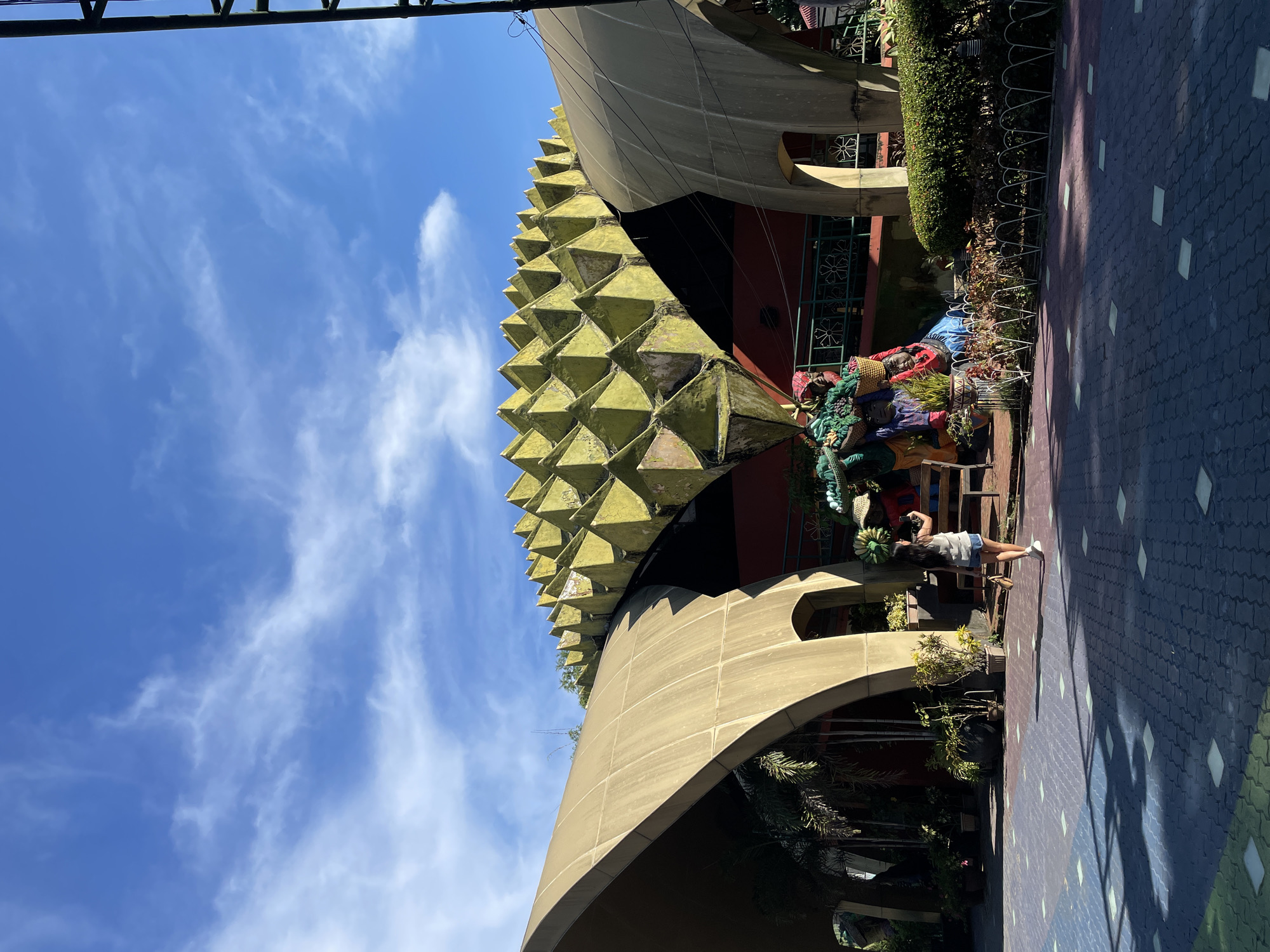 The image shows the durian-shaped building of the Durian Garden Restaurant in Davao City, Philippines. The building has a unique architectural design, with a spiky, yellow roof that resembles a durian fruit. The facade is adorned with curved concrete structures and arches, and a person is seen walking on the patterned ground in front of it. The sky is blue with scattered white clouds, and the area is decorated with plants and statues.