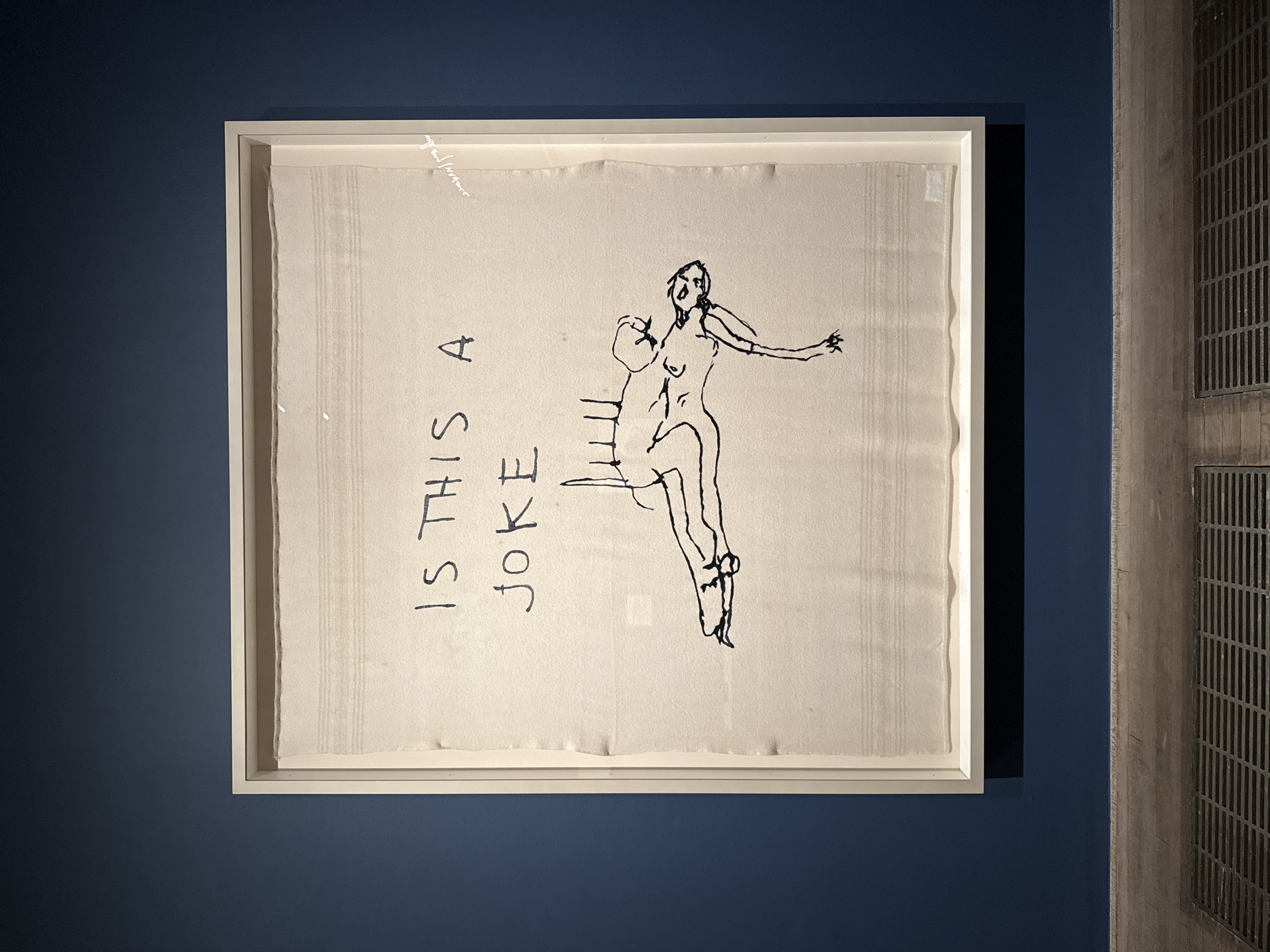 A framed artwork hangs on a dark blue wall. The artwork is a drawing on what appears to be a textured fabric or aged paper. The drawing depicts a figure seated on a chair, with one arm extended and an expression of distress or surprise. Scrawled text, "IS THIS A JOKE" is visible to the side of the figure. To the right of the artwork, a grated concrete wall structure is visible. The location is likely an art gallery or museum exhibiting contemporary art in London, United Kingdom.
