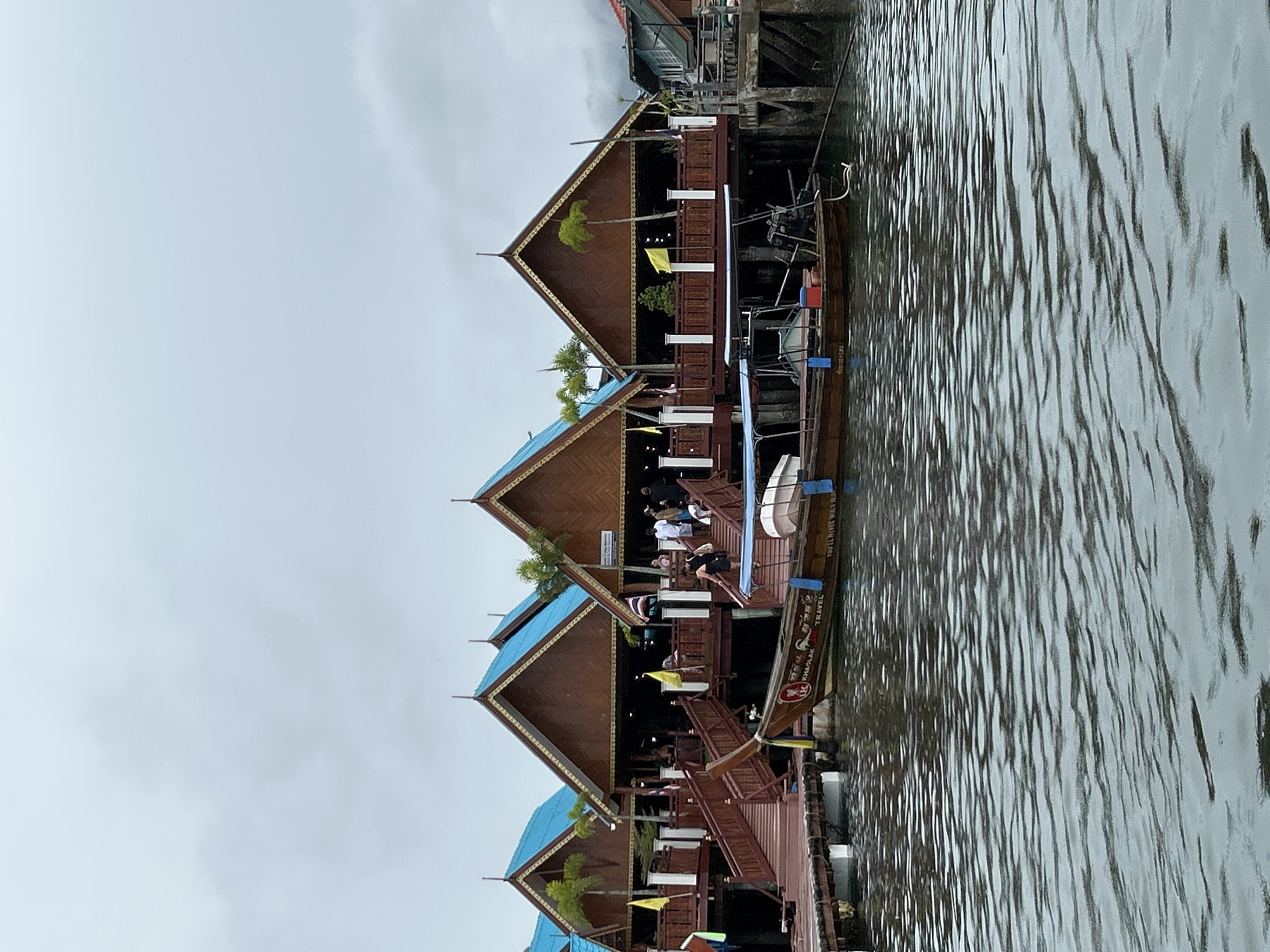 The image depicts a stilted village with several traditional wooden buildings featuring blue-tiled roofs and ornate trim. These structures are built on stilts over the water, and wooden walkways with railings connect them. A longtail boat is moored in the foreground, its wooden hull visible. Several people are seen walking on the walkways and stairs leading to the buildings. The sky is overcast, and the water is calm with visible ripples. The scene is identifiable as Ko Panyi, a floating village in Phang Nga Province, Thailand, known for its Muslim fishing community.