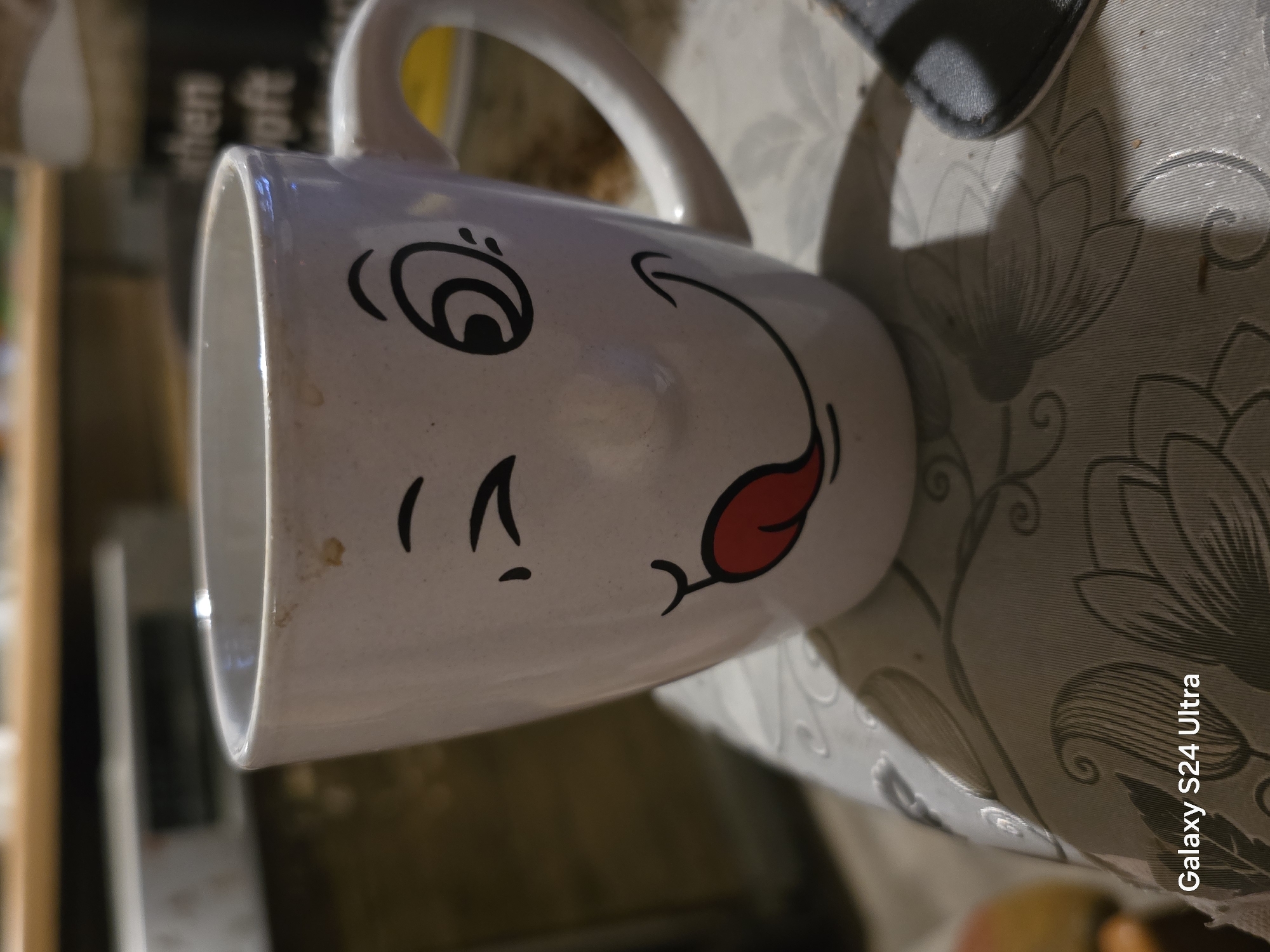 A close-up shot focuses on a white ceramic mug with a cartoon face drawn on it. The face features a winking eye, a large red tongue sticking out, and a smiling mouth. The mug is positioned on a table covered with a patterned tablecloth. In the background, a blurry shelf or counter is visible with some indistinct objects. The lighting is dim, casting shadows. The bottom left corner of the image displays "Galaxy S24 Ultra" in white text. There is no indication of the city of Göttingen, Germany, or any specific location within it.