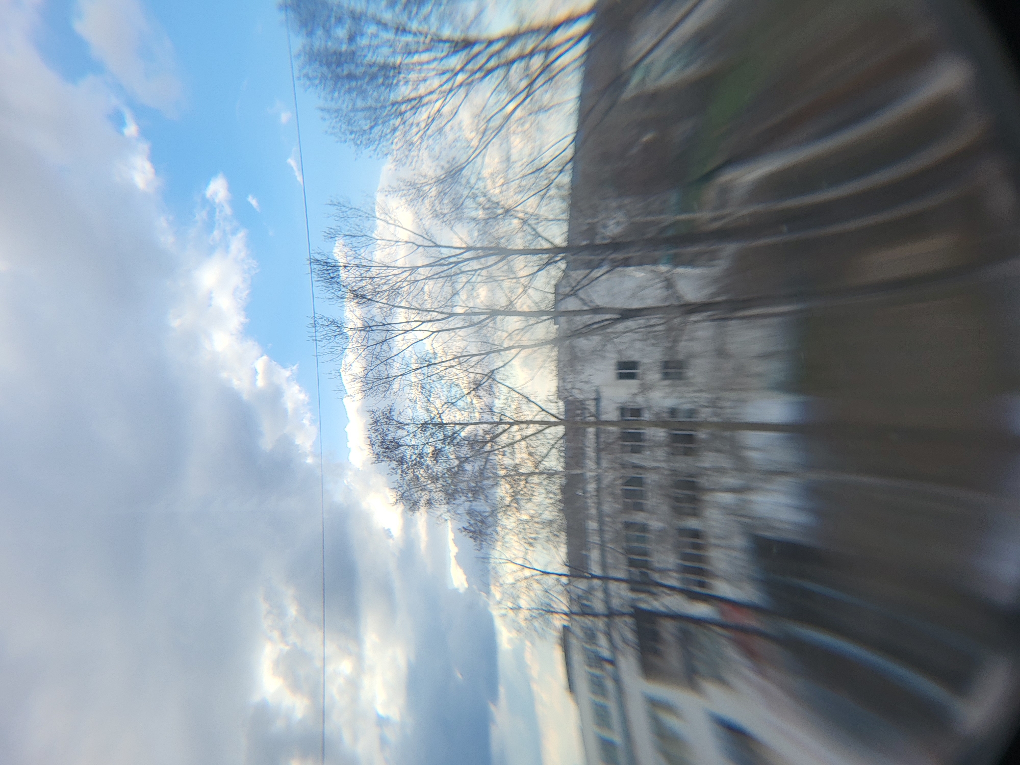 The image, possibly taken through a fish-eye lens, shows a distorted view of a building, trees, and a cloudy sky. The building appears to be a multi-story structure with multiple windows. Bare tree branches are visible in front of the building and against the backdrop of the sky. The sky is filled with clouds, with patches of blue visible.
Without more information about the location, it is difficult to pinpoint the exact location in the city of Chaikovsky, Russia.
