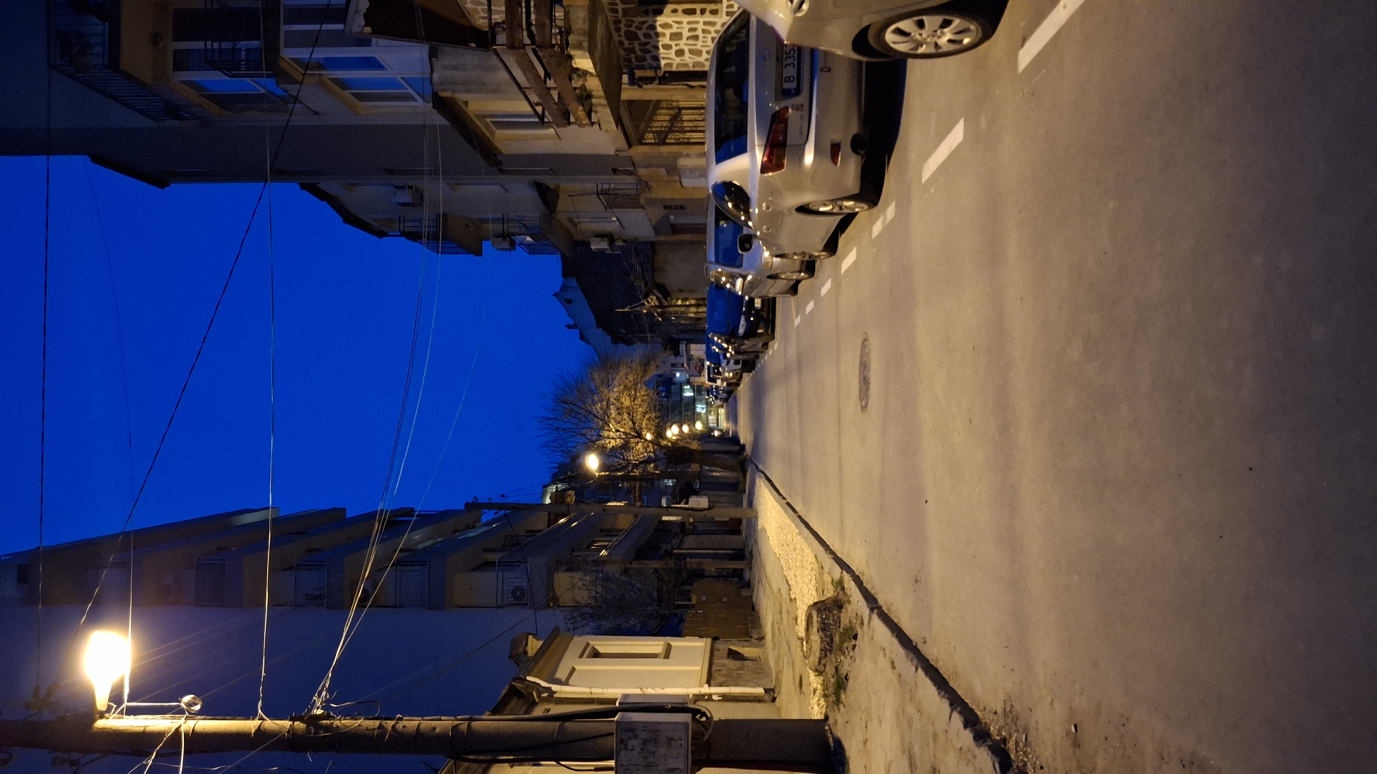 The image depicts a street in Varna, Bulgaria, at night. The street is lined with buildings of varying heights and architectural styles. Parked cars are visible along the right side of the street. Streetlights illuminate the scene, casting shadows and creating a contrast between light and dark areas. Power lines are visible above the street. The sky is a dark blue. There are no people visible in the image.