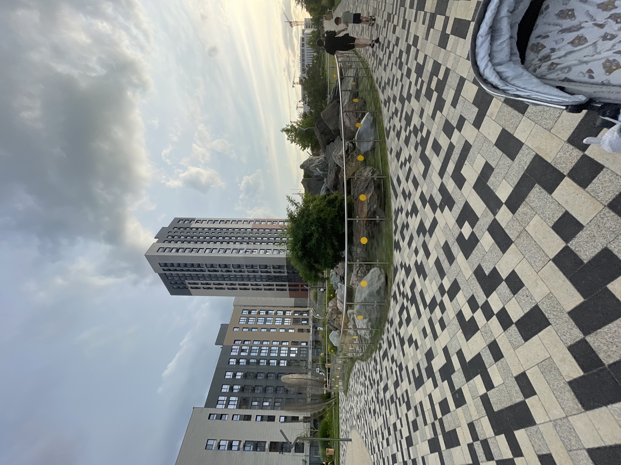 In the city of Stavropol, Russia, an outdoor scene unfolds with a striking geometric paved walkway in the foreground, featuring a mosaic of black and light-colored square tiles laid in a dynamic pattern. The walkway leads toward a landscaped area with large rocks and dense greenery, enclosed by a sleek metal railing with yellow circular accents. Behind this area, several modern multi-story apartment buildings rise, their facades characterized by numerous windows and a contemporary architectural style. In the middle distance, a tall, slender skyscraper stands out, dwarfing its neighboring structures. Further in the background, construction cranes are visible, suggesting ongoing development in the area. Two individuals, a man and a child, are walking away from the camera along the paved path, their silhouettes outlined against the slightly overcast sky. A baby stroller is partially visible in the lower right corner of the frame. The overall atmosphere is one of urban development and outdoor recreation.