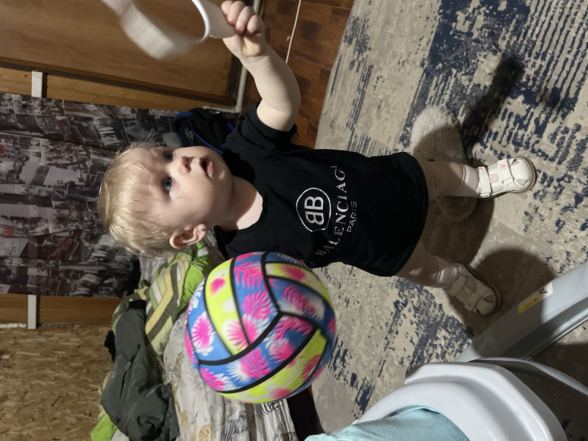 A toddler stands indoors, holding a white plastic object in their right hand. The child has blonde hair and blue eyes, and is wearing a black t-shirt with a "Balenciaga Paris" logo, pink socks, and white sandals. They are also positioned near a colorful beach ball. The environment includes a rug, a door, and patterned bedding. A baby walker is visible in the lower right corner. Based on the environment, the location appears to be indoors, potentially a bedroom or living space. The city of Vesyoloye, Russia, cannot be confirmed based solely on the image.