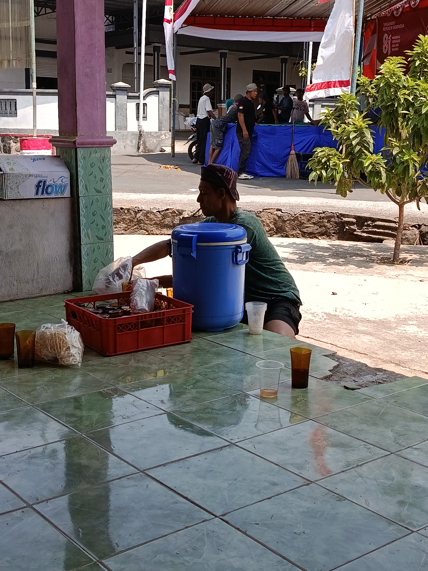 In Jurug, Indonesia, a man wearing a green t-shirt and black shorts is seen sitting on the ground next to a large blue insulated container. He is reaching into a plastic bag, holding a plastic cup, as he retrieves items from a red plastic crate filled with more plastic cups and what appear to be snacks. The crate also contains several plastic-wrapped items. The scene unfolds on a tiled floor, with several other cups and a plastic bag of snacks scattered nearby. In the background, a white tent with red and white bunting suggests an outdoor event. Several people are visible behind a blue tarp, some interacting and one appearing to push. A tree with green foliage grows to the right of the frame. The environment suggests a hot, sunny day.