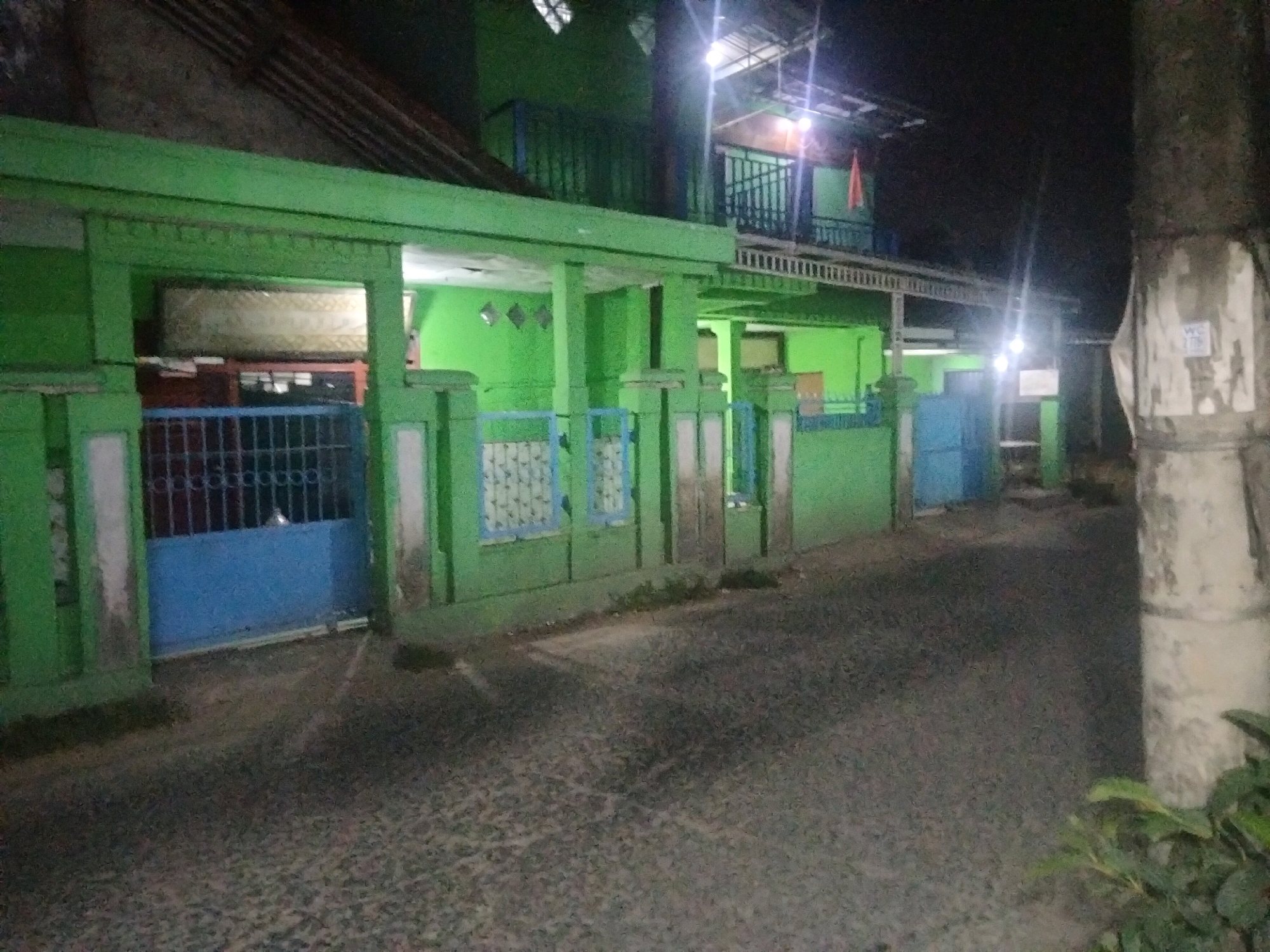 The image depicts a nighttime scene on a street in Suka Sari, Indonesia. A bright green, two-story building with a tiled roof dominates the left side of the frame. The building has balconies on the upper floor, some with metal railings. Various doors and windows are visible, some with decorative elements. A blue gate is positioned on the ground floor. The street in front of the building is paved with what appears to be cobblestones or similar textured material and is illuminated by streetlights. A large tree trunk, partially covered in what looks like plaster or concrete, stands on the right side of the image, with some foliage visible at its base. The overall atmosphere is dimly lit, characteristic of nighttime in a residential or commercial area. There are no people clearly visible in the image.