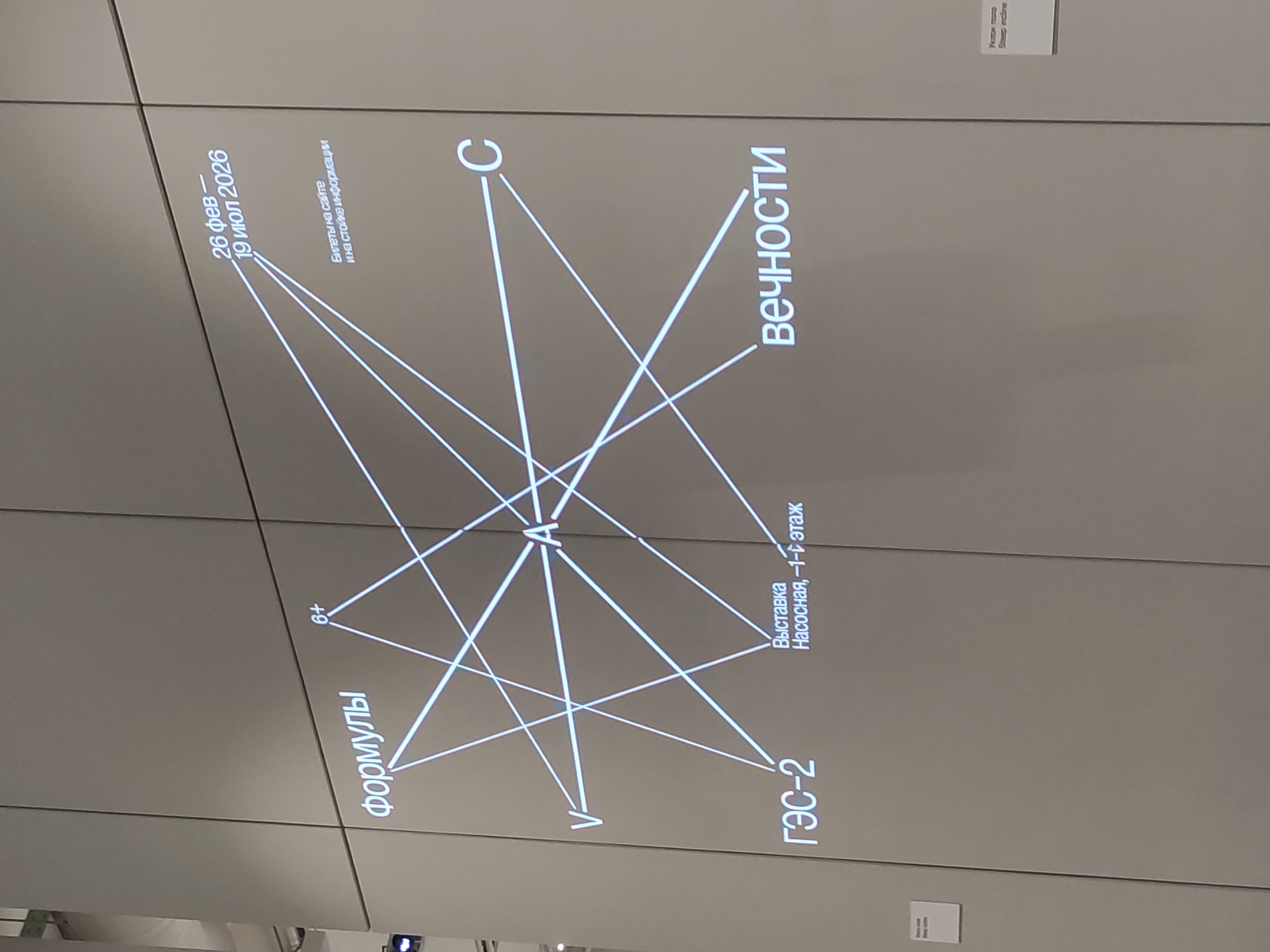 Here is a forensic description of the image.
The image shows a large, wall-mounted display featuring white text and lines against a gray background, seemingly part of an exhibition or informational installation. The text includes the following: "Формулы" (Formulas), "6+", "26 фев - 19 июл 2026" (26 Feb - 19 Jul 2026), "Билеты на сайте и на стойке информации" (Tickets available on the website and at the information desk), "ГЭС-2" (GES-2), "Выставка Насосная, -1-й этаж" (Pumping Station Exhibition, -1st floor), and "Вечности" (Eternity), along with the letters 'V', 'A', and 'C'. Lines connect these words and letters, creating a complex network.
Smaller, unreadable text and labels are visible near the lower and upper right corners of the display. The display appears to be made up of multiple panels. The lighting in the room is somewhat dim, causing shadows on the display.
There is no identifiable location or city information in the image.