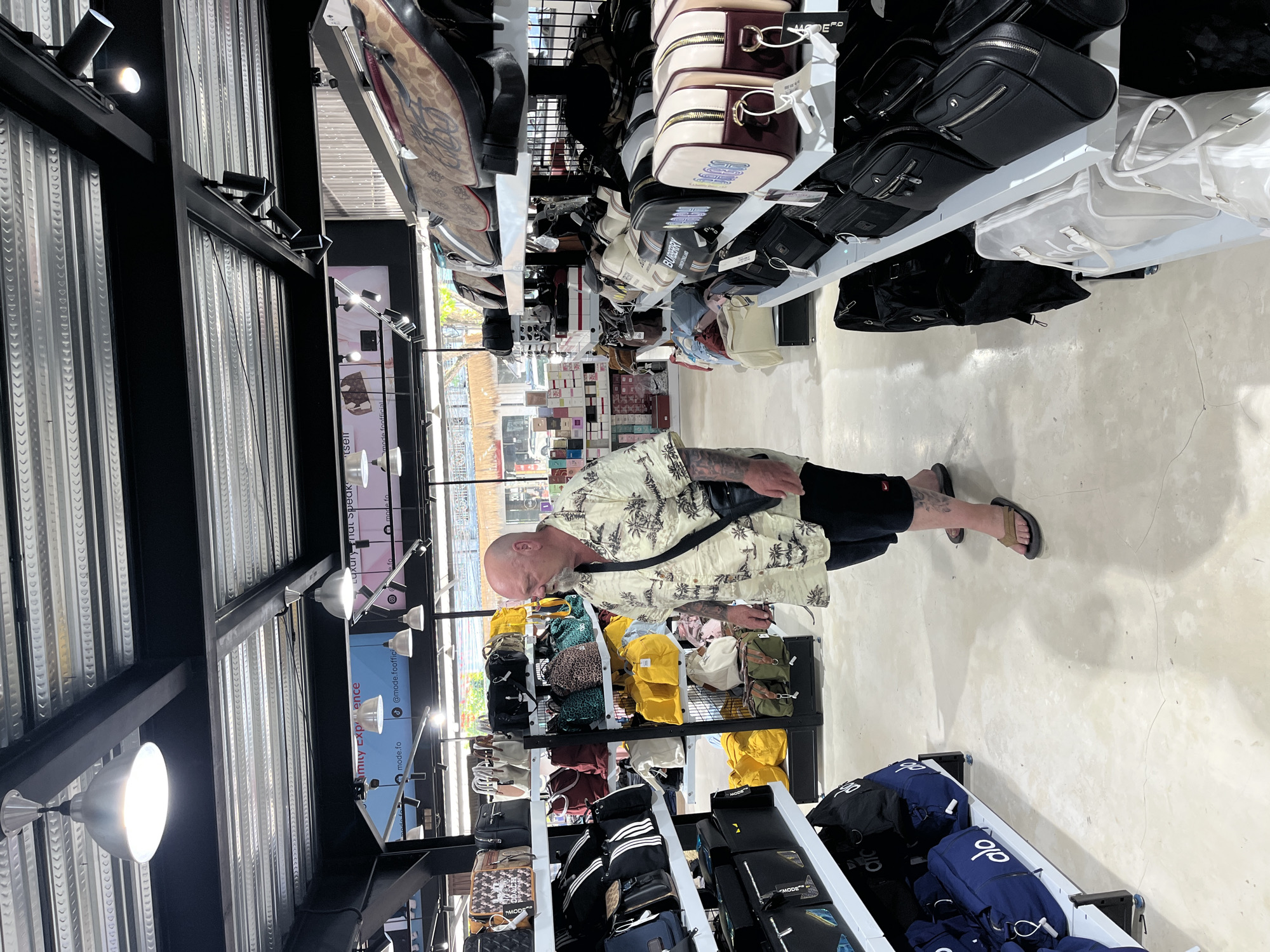 In the city of North Kuta, Indonesia, a bald, middle-aged man with tattoos on his arms is browsing a store that sells bags and luggage. He is wearing a tropical-print short-sleeved shirt, black shorts, and brown flip-flops. He is carrying a black crossbody bag. The store has rows of shelves displaying various types of bags, including duffel bags, handbags, and shoulder bags, in different colors and designs. The lighting is bright, with fluorescent lights visible on the ceiling. The floor is a light-colored, polished concrete. The store appears to be well-stocked, with a wide selection of merchandise.