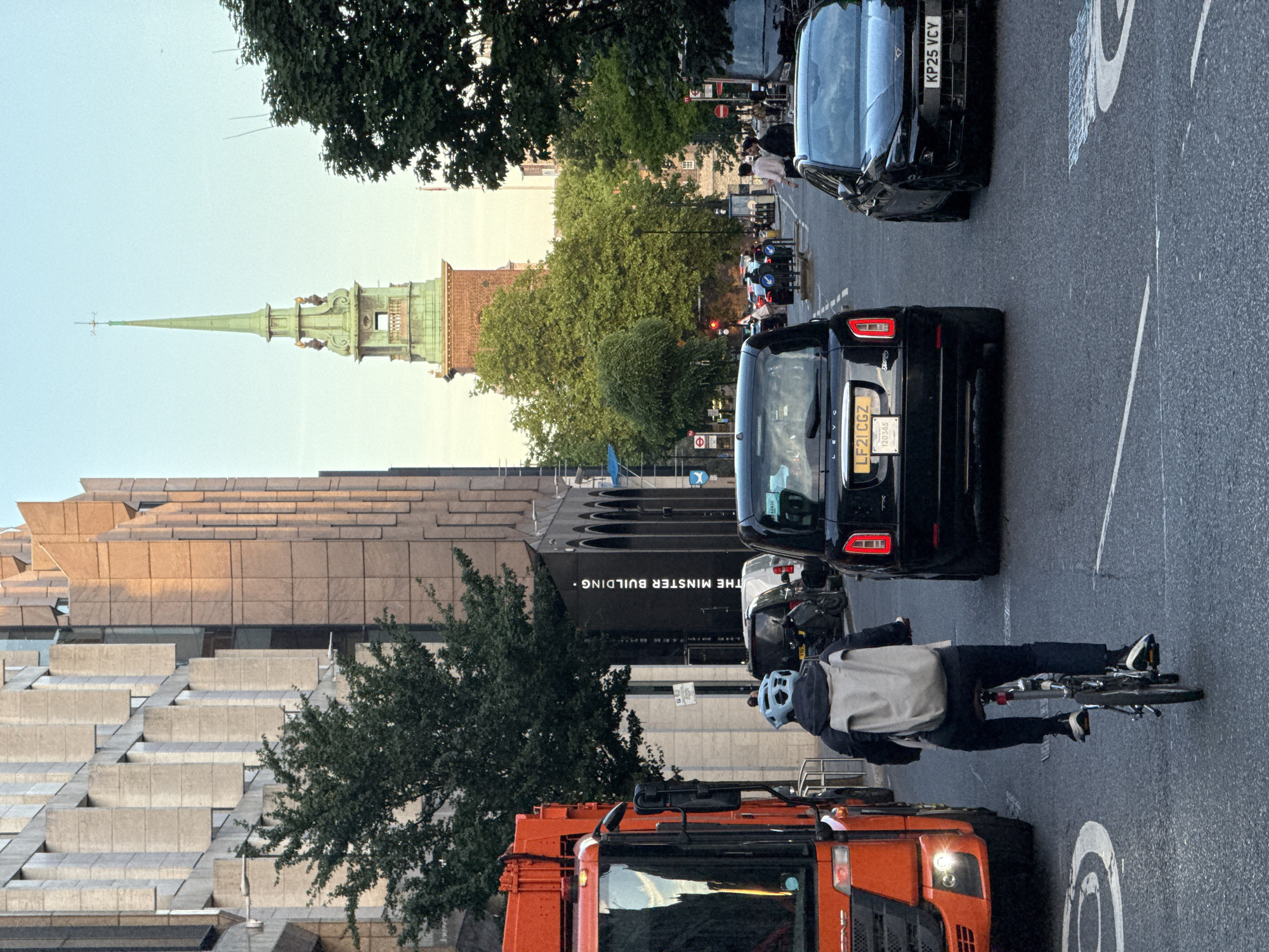 The image depicts a street scene in London, United Kingdom. The focus is on the road with a cyclist wearing a blue helmet, grey jacket and dark trousers. The cyclist is positioned in front of an orange truck. Two black cars are visible in the background, partially obscuring the view of pedestrians and a green spire in the distance. A building with a terraced, stepped design is to the left of the frame, and a sign saying "The Minster Building" is also visible. The road is marked with white lines. Trees are visible in several locations.