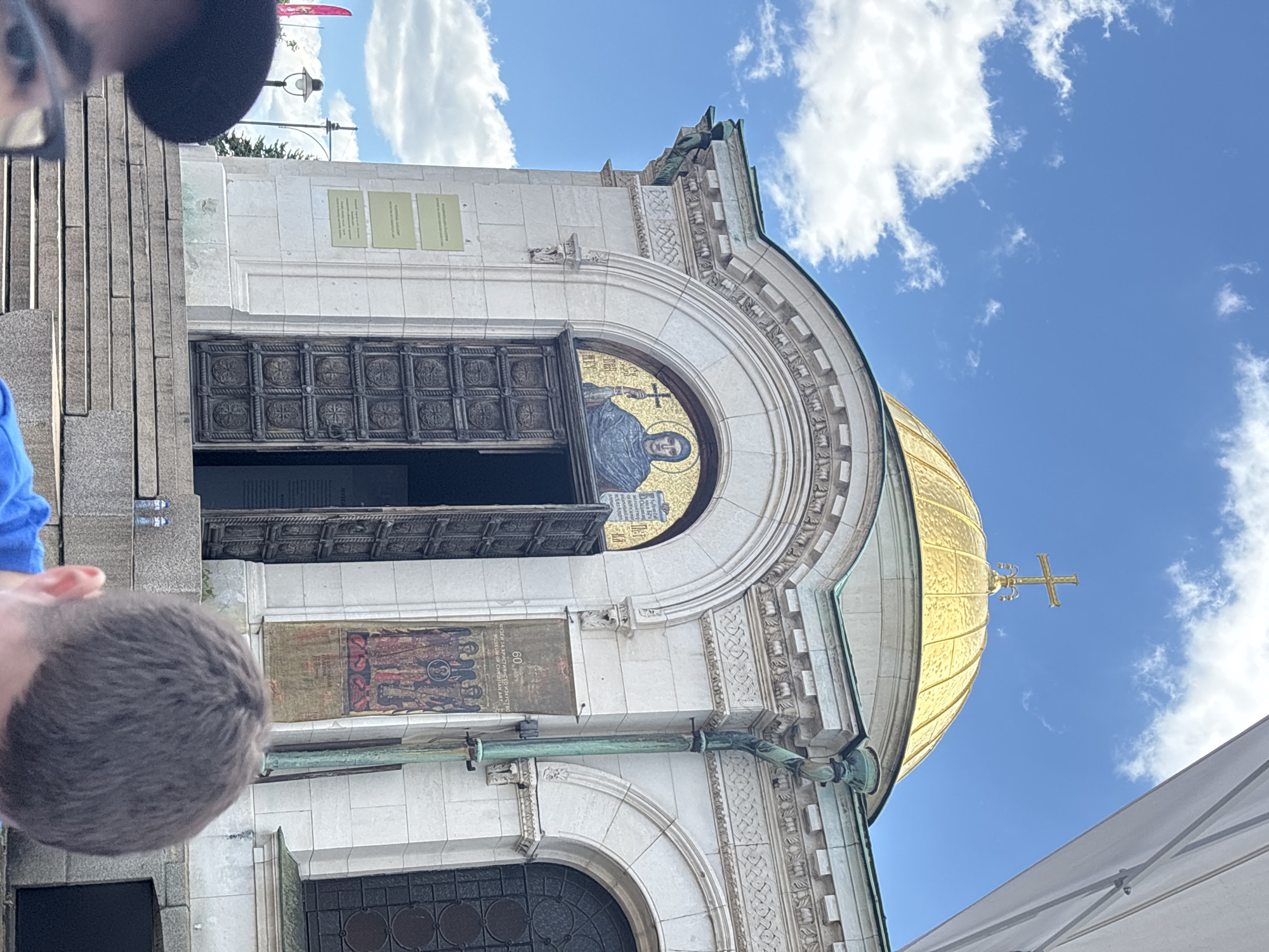 The image depicts the entrance and part of the golden dome of the St. Alexander Nevsky Cathedral in Sofia, Bulgaria. The entrance features a large, ornate wooden door that is partially open. Above the door, there is a mosaic depicting a saint. To the left of the door, plaques and paintings are visible. The dome is gold in color and adorned with a cross at its peak. The sky is blue with some white clouds. Parts of at least two people are visible in the foreground, including the top of their heads and partial clothing.