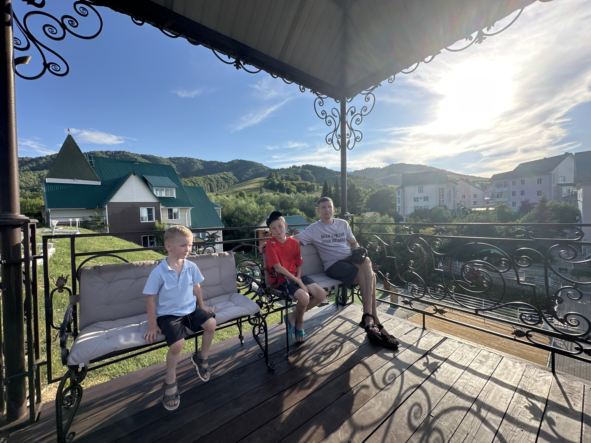 Three males are seated on a wrought-iron bench on a wooden porch. The male on the far left is a young boy wearing a light blue polo shirt and black shorts, with sandals on his feet. The middle male is a young boy wearing a red t-shirt and black shorts, with blue flip-flops. The male on the right is an adult wearing a gray t-shirt with text and black shorts, with sandals on his feet. The porch has a wooden floor and a decorative wrought-iron railing. Behind the porch, there are green rolling hills and buildings. The sky is blue with scattered white clouds, and the sun is shining brightly, casting shadows on the porch. The scene appears to be in a residential area in Даниловка (алтайский), Russia, likely a vacation or resort setting given the scenic background.