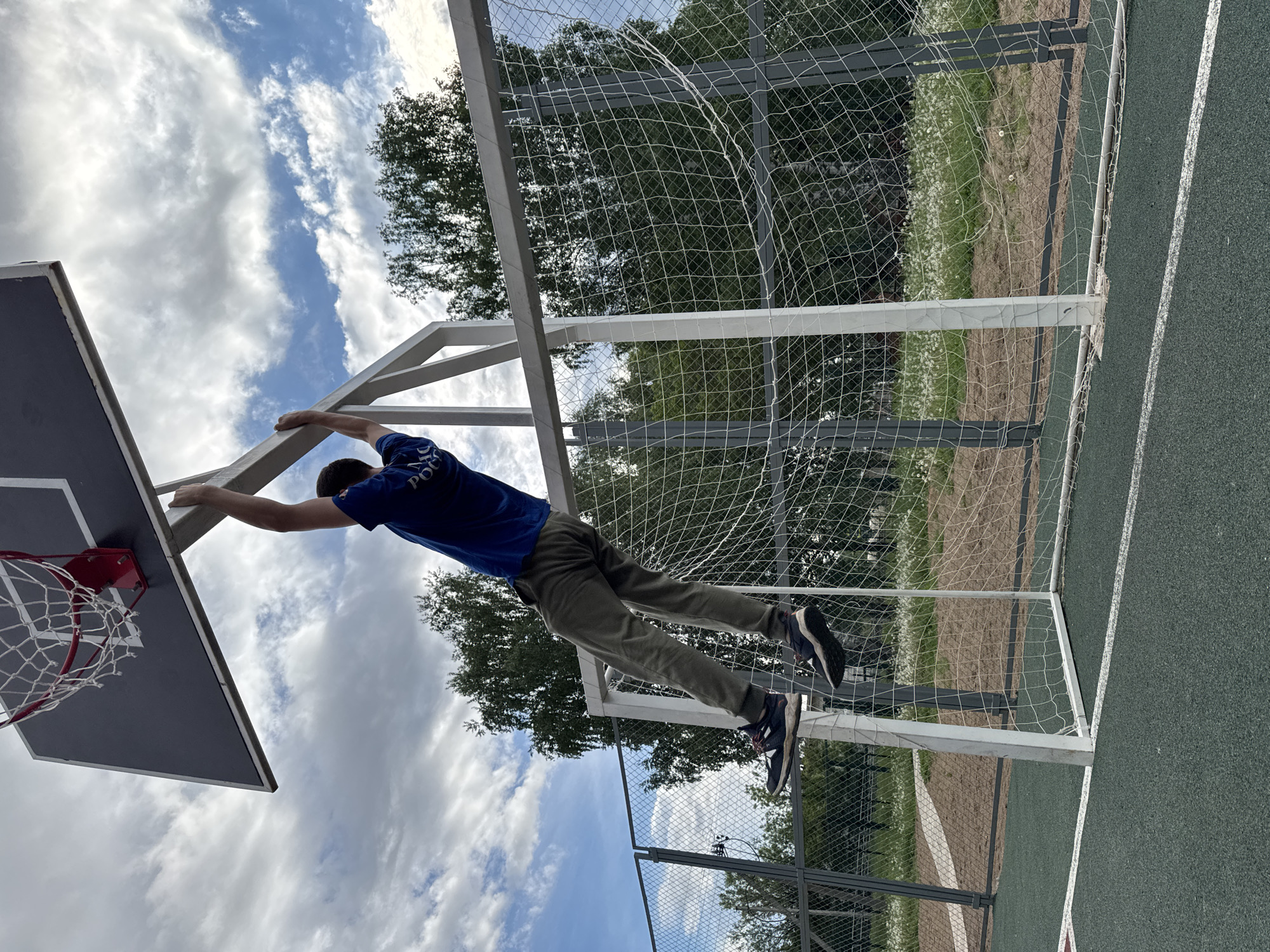 A young person hangs from a basketball hoop in Вассята, Russia. The person is wearing a blue shirt and gray pants. A soccer goal is also visible behind the basketball hoop. The sky is cloudy. The ground is a green artificial surface.