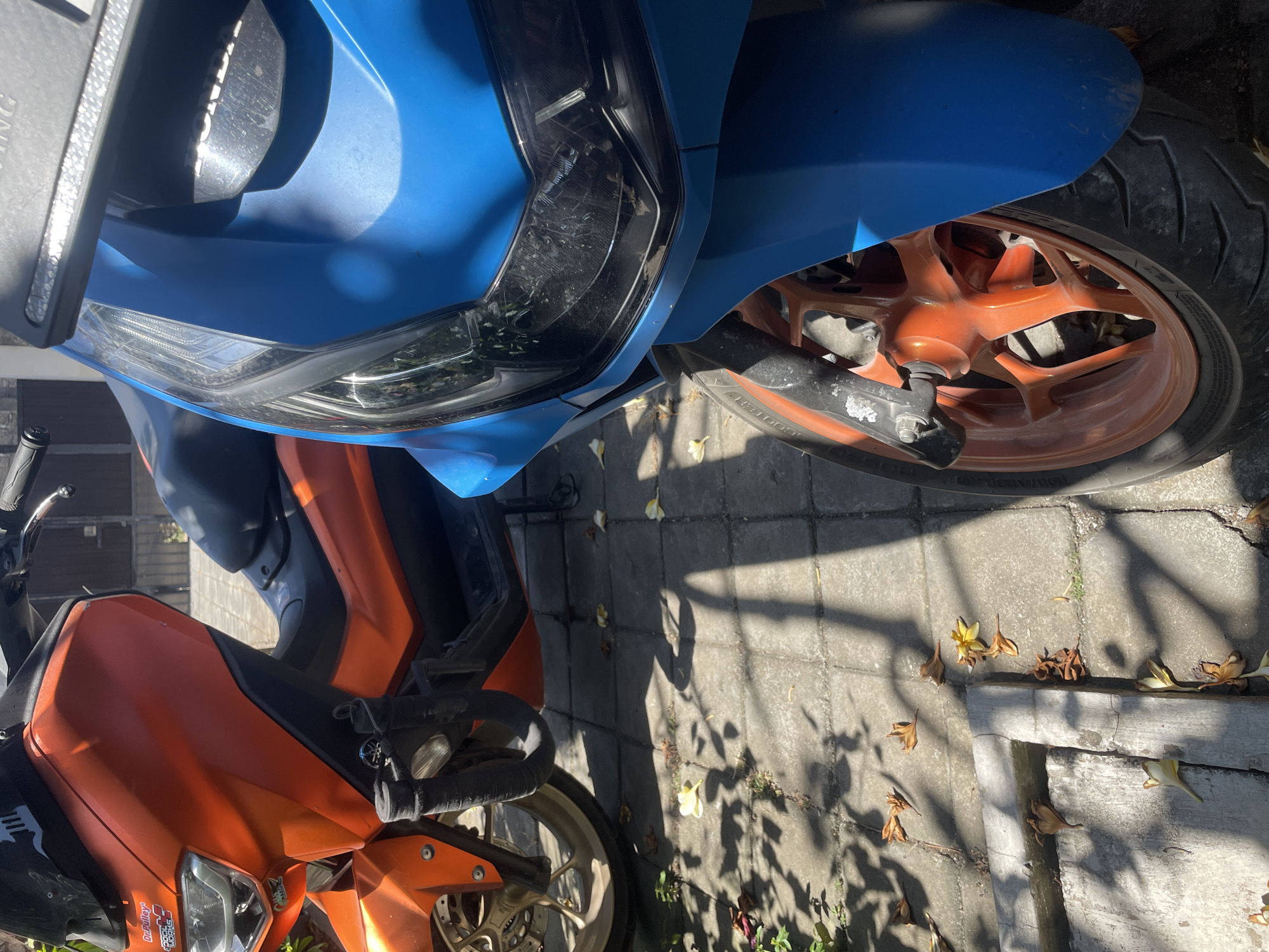 In Kuta Selatan, Indonesia, two scooters are parked on a sun-drenched, tiled pavement. The scooter on the left is orange, with a black windshield and black handlebars. A portion of its orange wheel is visible. The scooter on the right is predominantly blue, with a black headlight assembly and part of its front orange wheel visible. The ground is partially covered with fallen leaves, and shadows from overhead foliage cast dappled patterns.