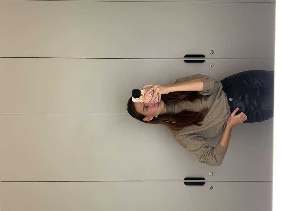 A female subject is depicted in a mirror selfie, captured from the waist up. She stands with her left hand resting on her hip and her right hand holding a smartphone, which partially obscures the lower portion of her face. The subject wears a light brown or beige long-sleeved turtleneck sweater and dark, lustrous high-waisted pants. Visible accessories include a long dangle earring on her left ear, a ring on her left ring finger, and a ring on her right middle finger. The smartphone case is light-colored and features multiple small, stylized motifs. The background, reflected in the mirror, consists of a flat, light grey or beige wall composed of several vertical panels resembling closet or changing room doors. Two distinct black vertical handles and two circular keyholes are visible on two adjacent panels. The lighting is uniform, suggesting an indoor setting such as a retail changing room, a fitting room, or a private closet area. The specific location within the city of Krasnoyarsk, Russia, cannot be determined from the visual information provided in the image.
