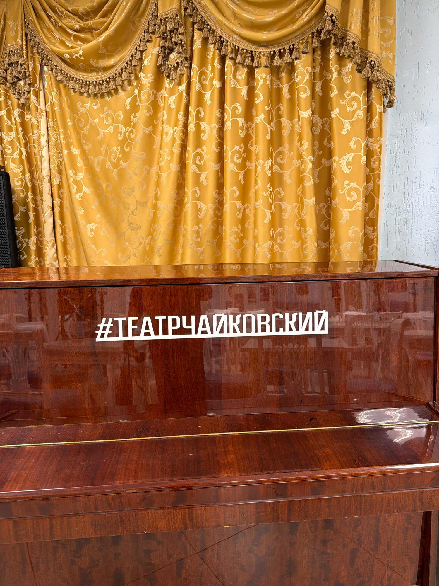 The image shows a close-up of a polished dark wood piano, likely located in the city of Chaikovsky, Russia. A white hashtag symbol followed by the Cyrillic text "#ТЕАТРЧАЙКОВСКИЙ" is affixed to the front surface of the piano. Behind the piano, a decorative yellow curtain with a damask-like pattern and tassels is visible. The curtain is draped and appears to be part of a stage or performance setting. The piano's surface is reflective, showing distorted images of chairs and the curtain. The overall scene suggests an indoor location, possibly a theater or concert hall in Chaikovsky.