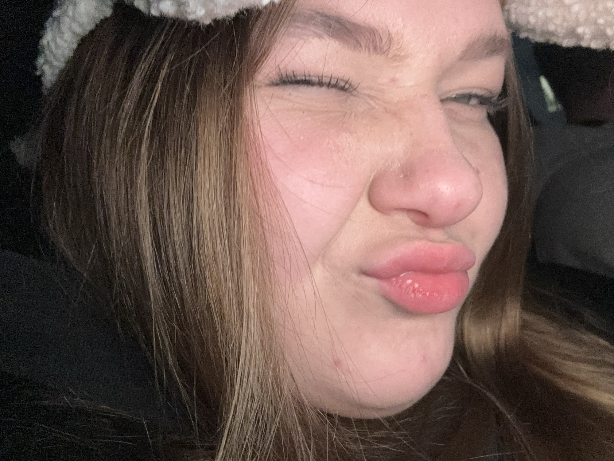 The image shows a close-up of a woman's face. She is winking one eye and making a "duck face" with her lips. Her hair is brown with blonde highlights, and she is wearing a fuzzy white headband. Her skin is fair and she has freckles on her nose. The background is dark and out of focus, but it appears to be the interior of a car. There is no information in the image to suggest a location in Chaikovsky, Russia.